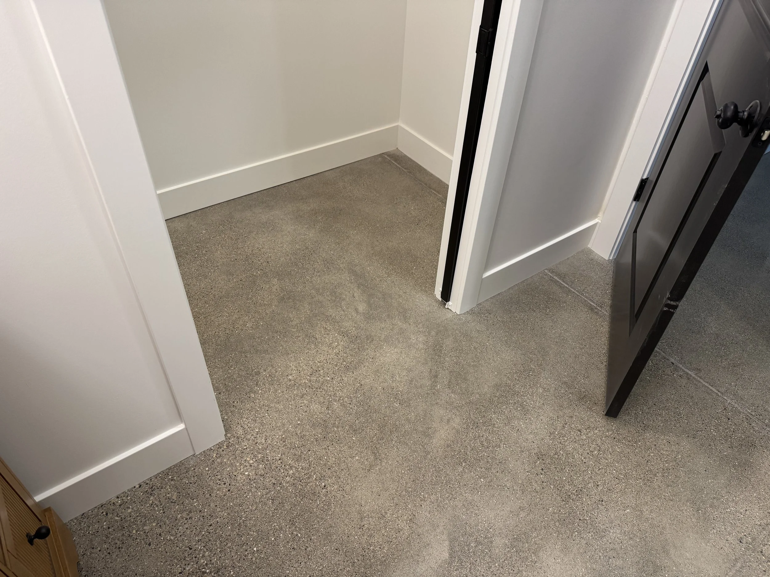 Corner of a room with gray textured concrete flooring, white walls with white baseboards, and a partially open black door.