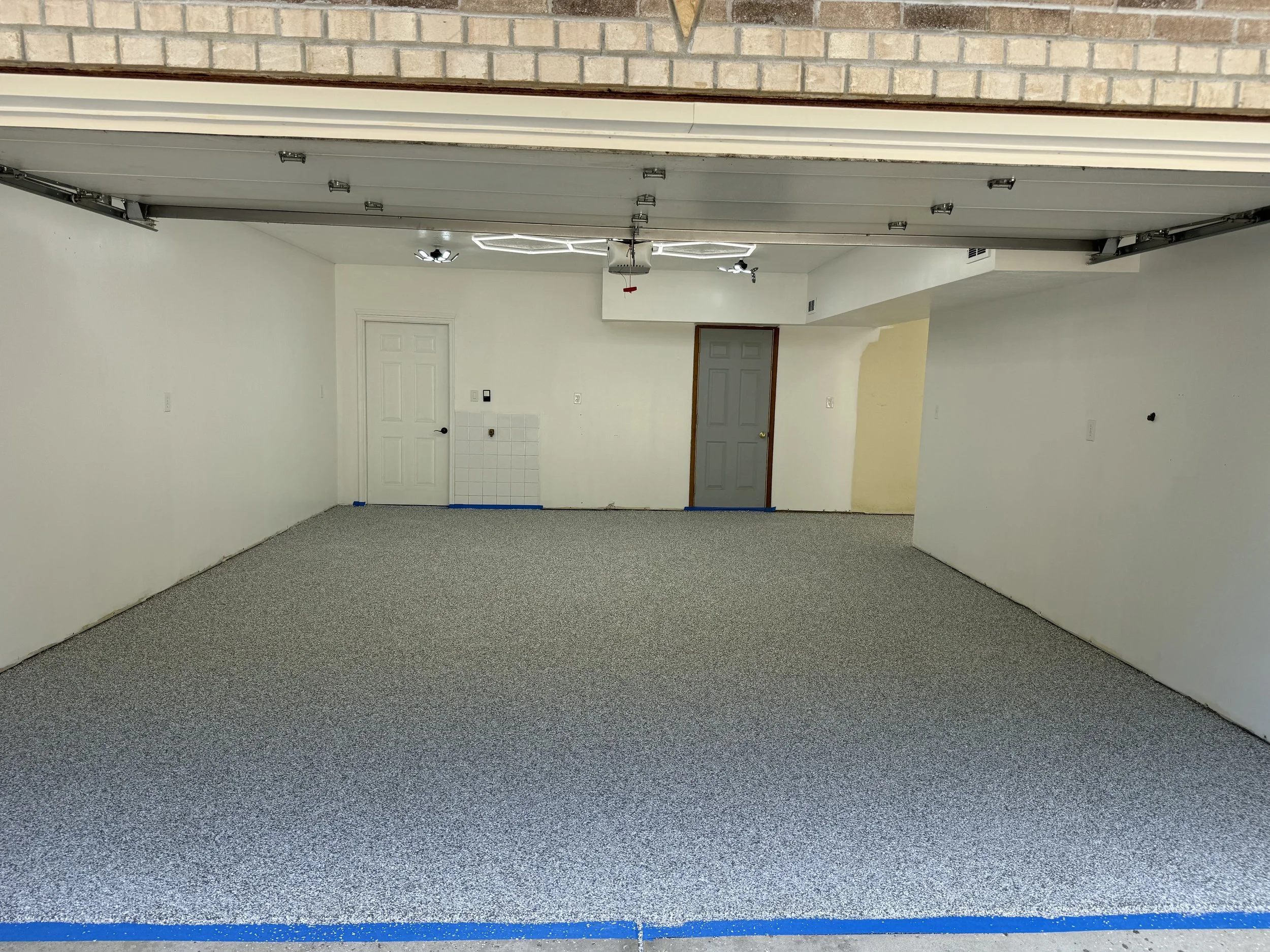 Empty garage with carpeted floor, two closed doors, white walls, and an open garage door showing a brick exterior.