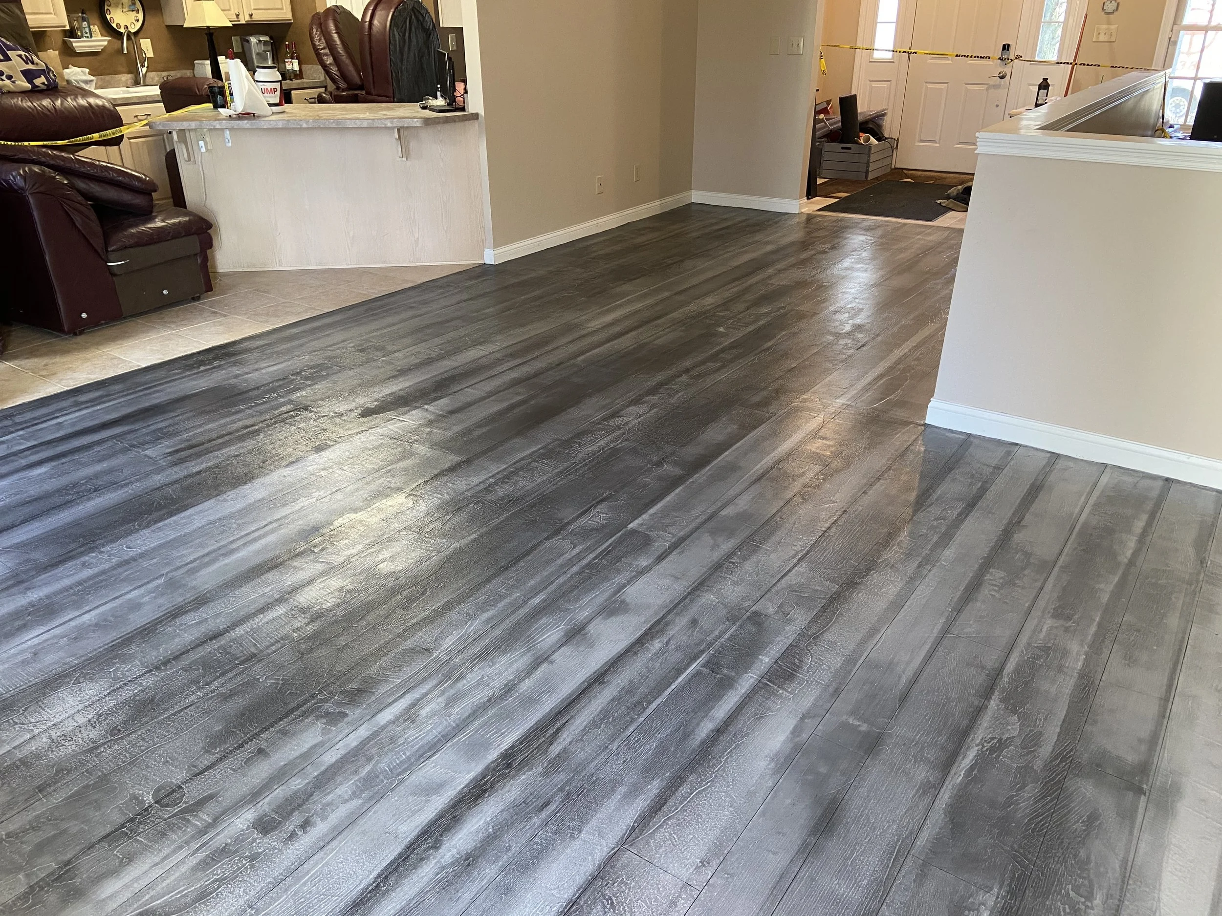Interior of a living space with dark wood flooring, beige walls, and a kitchen area in the background with a brown leather chair. Some caution tape is visible near the entrance, indicating area restriction.