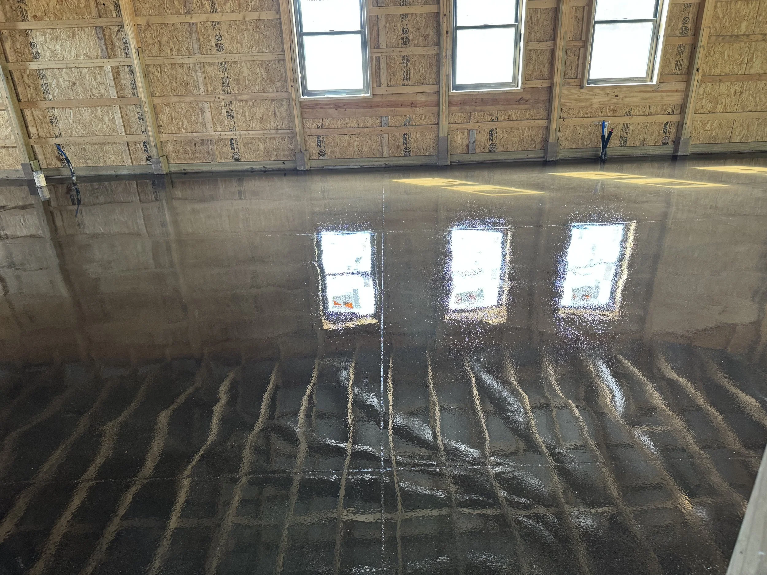 Interior of a building under construction with a shiny, wet concrete floor reflecting windows and unfinished wooden walls.