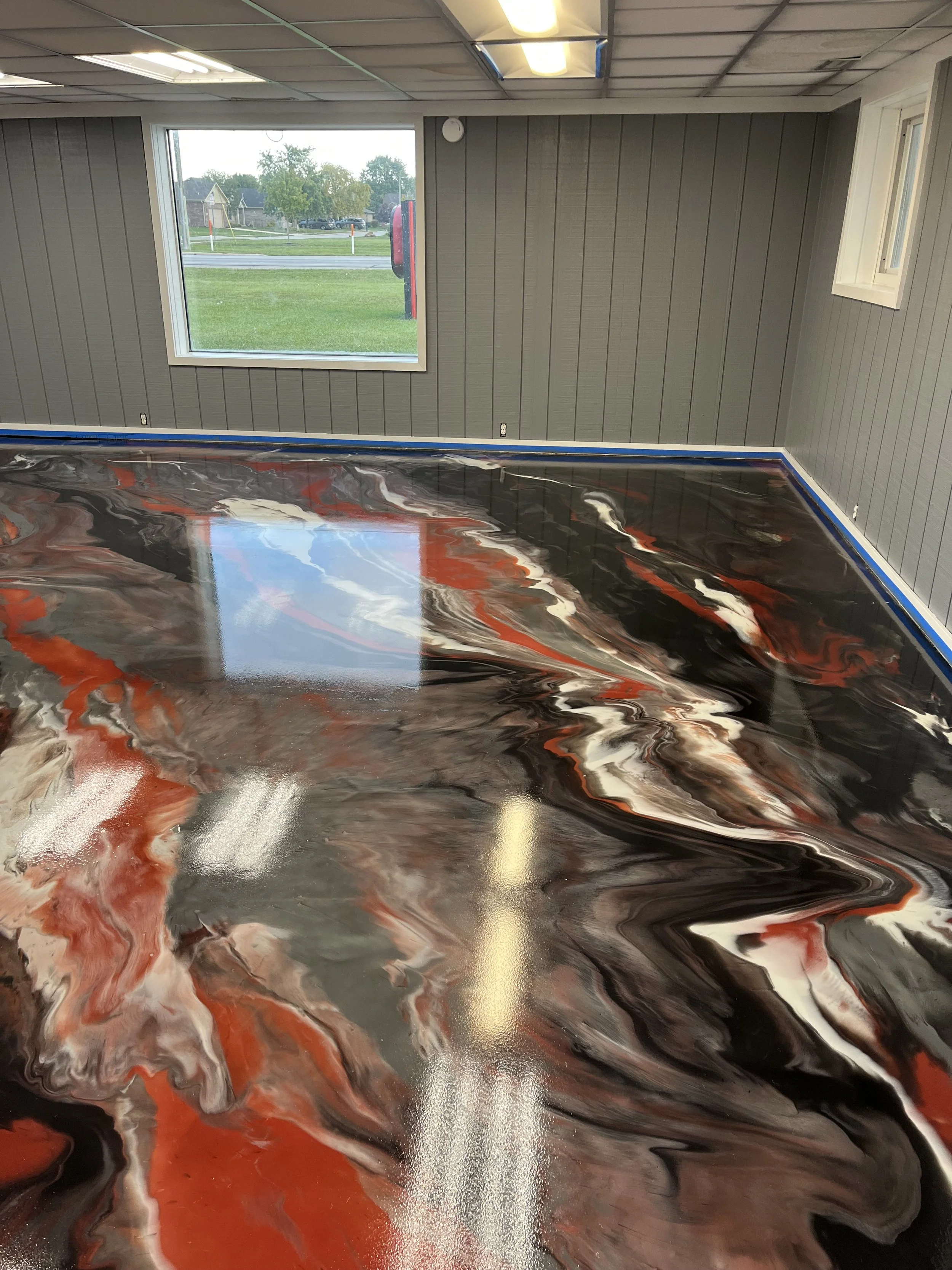 Interior room with marbled black, white, orange, and brown epoxy flooring, gray paneled walls, a large window showing a green outdoor area, and a reflection of ceiling lights on the shiny floor.