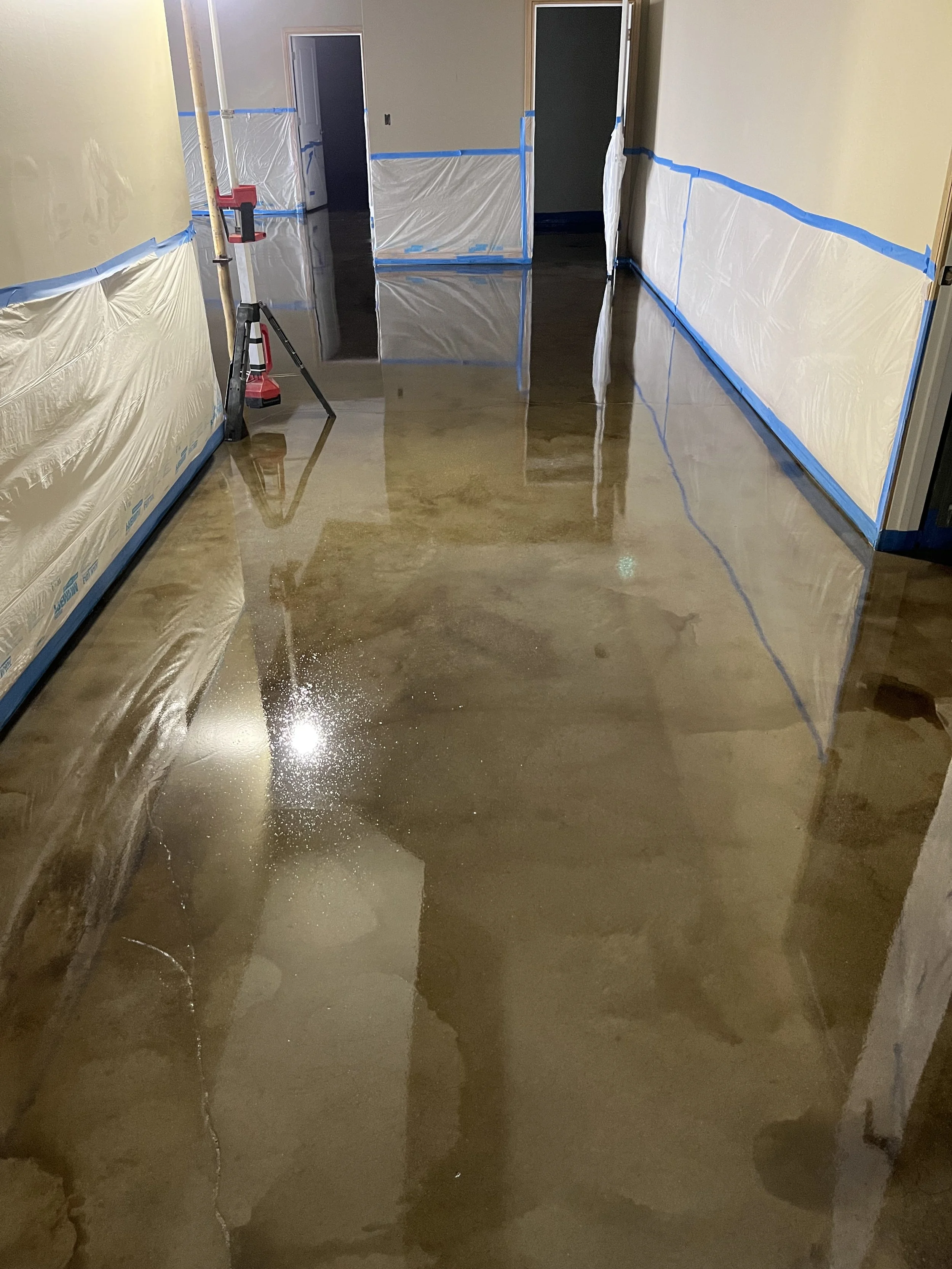 Indoor hallway with a freshly polished, shiny concrete floor and walls covered with painter's plastic and blue painter's tape, indicating ongoing renovation or painting.
