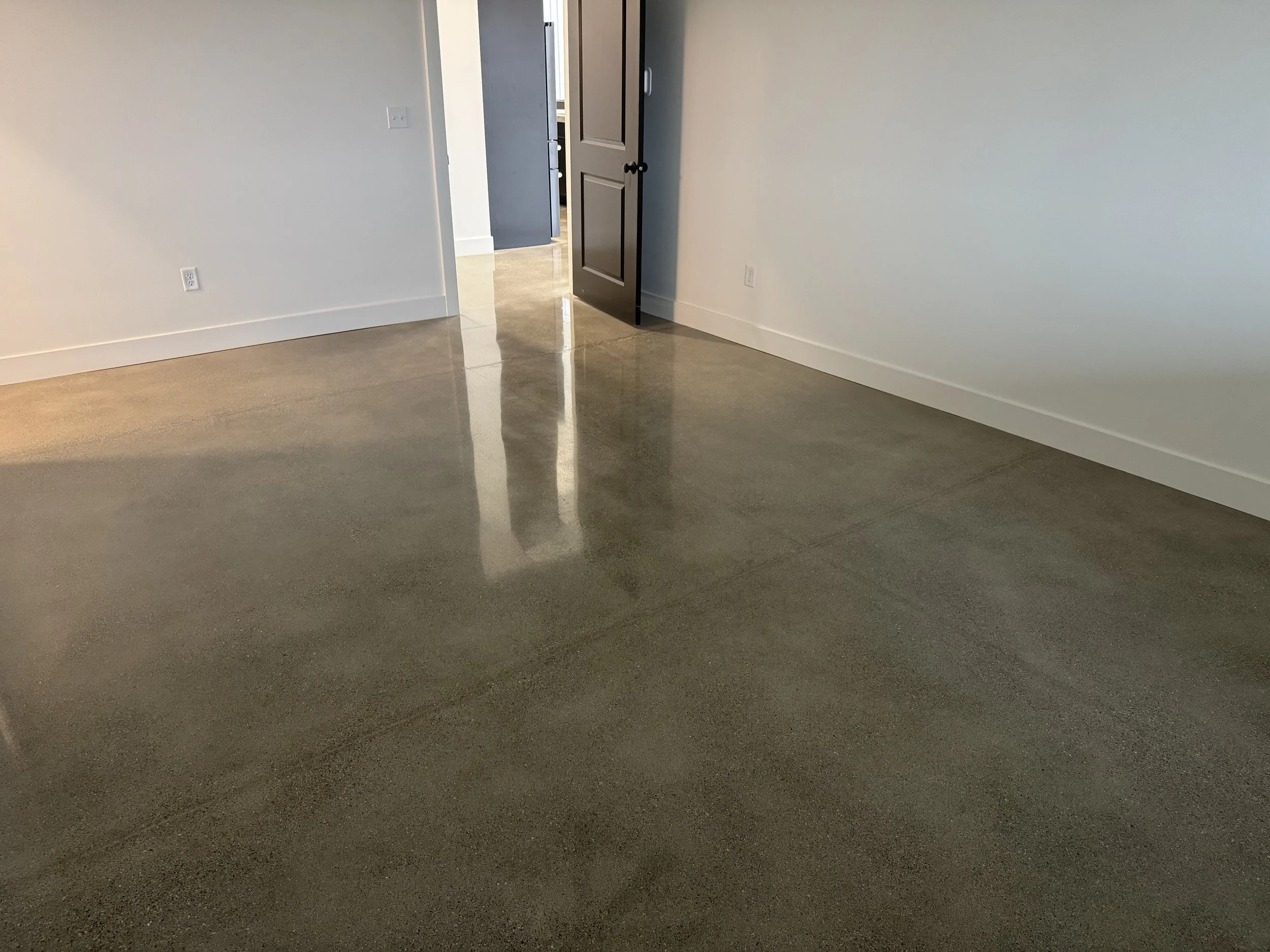 Empty room with polished concrete floor, white walls, and white baseboards. There is a partially open dark brown door and a wall outlet visible.