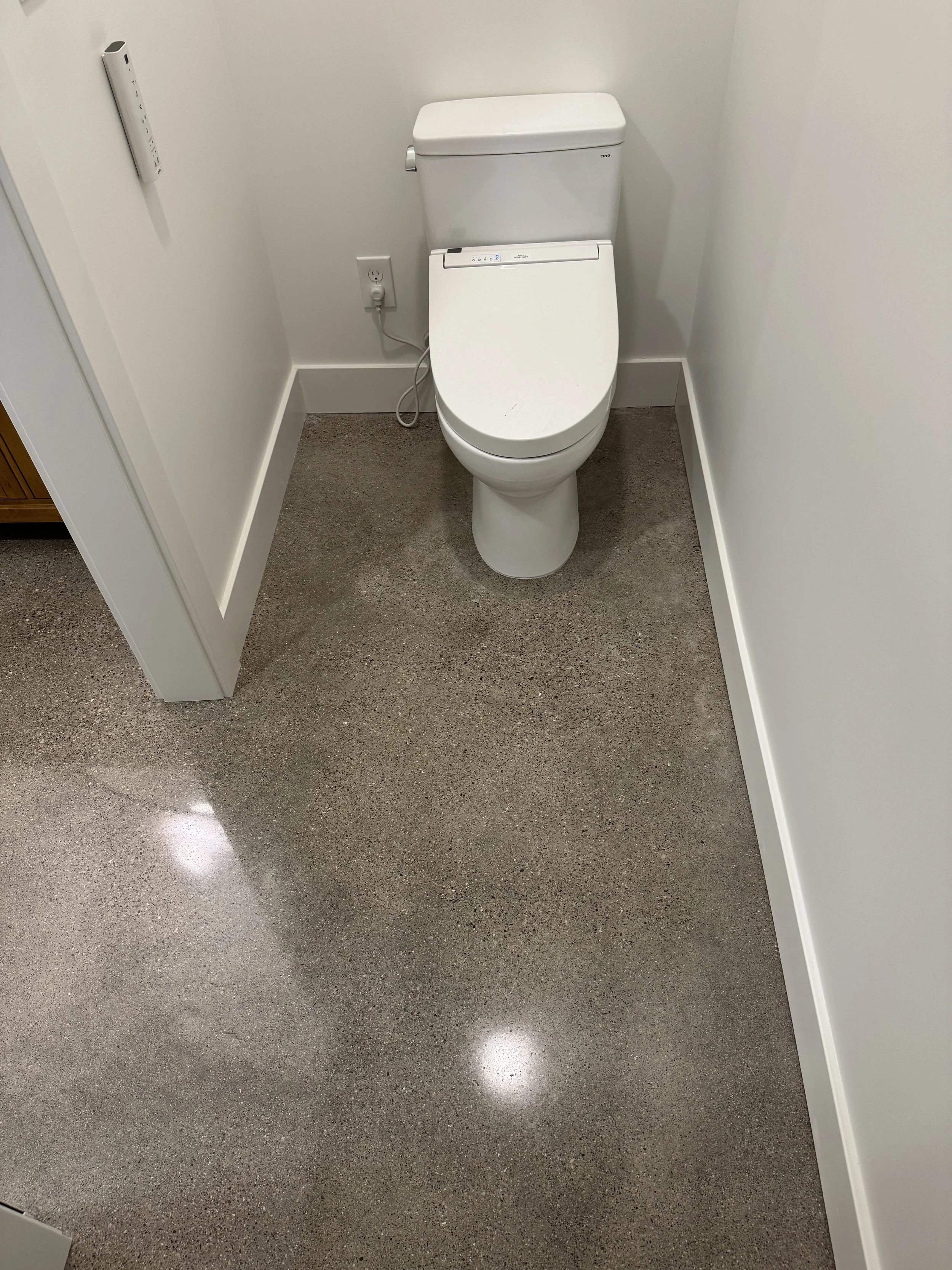 A modern toilet in a small bathroom with polished concrete flooring and plain white walls.