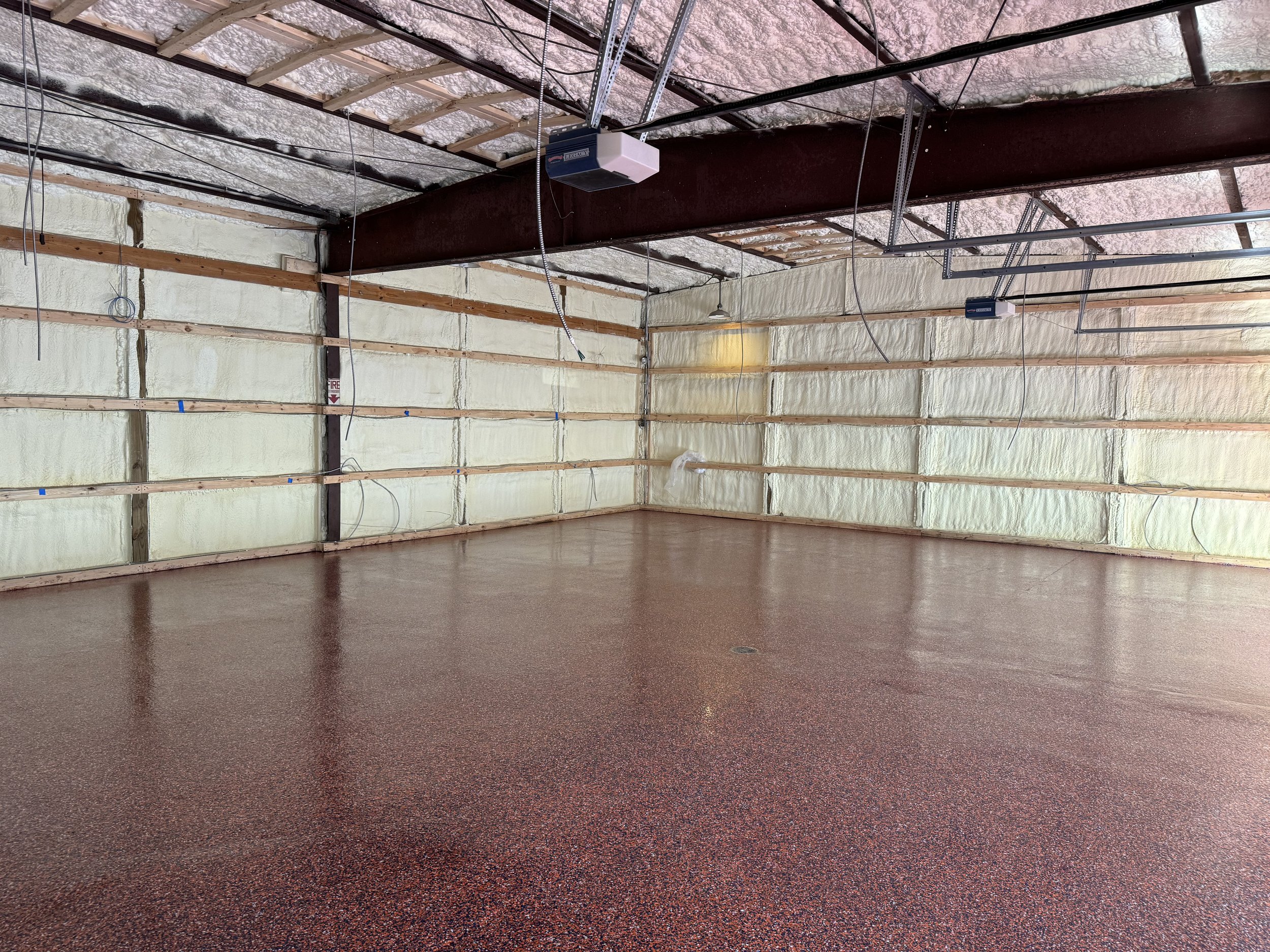 Empty garage with spray foam insulation on the walls and ceiling, and a red speckled epoxy floor. The ceiling shows exposed beams, insulation, and some wiring.