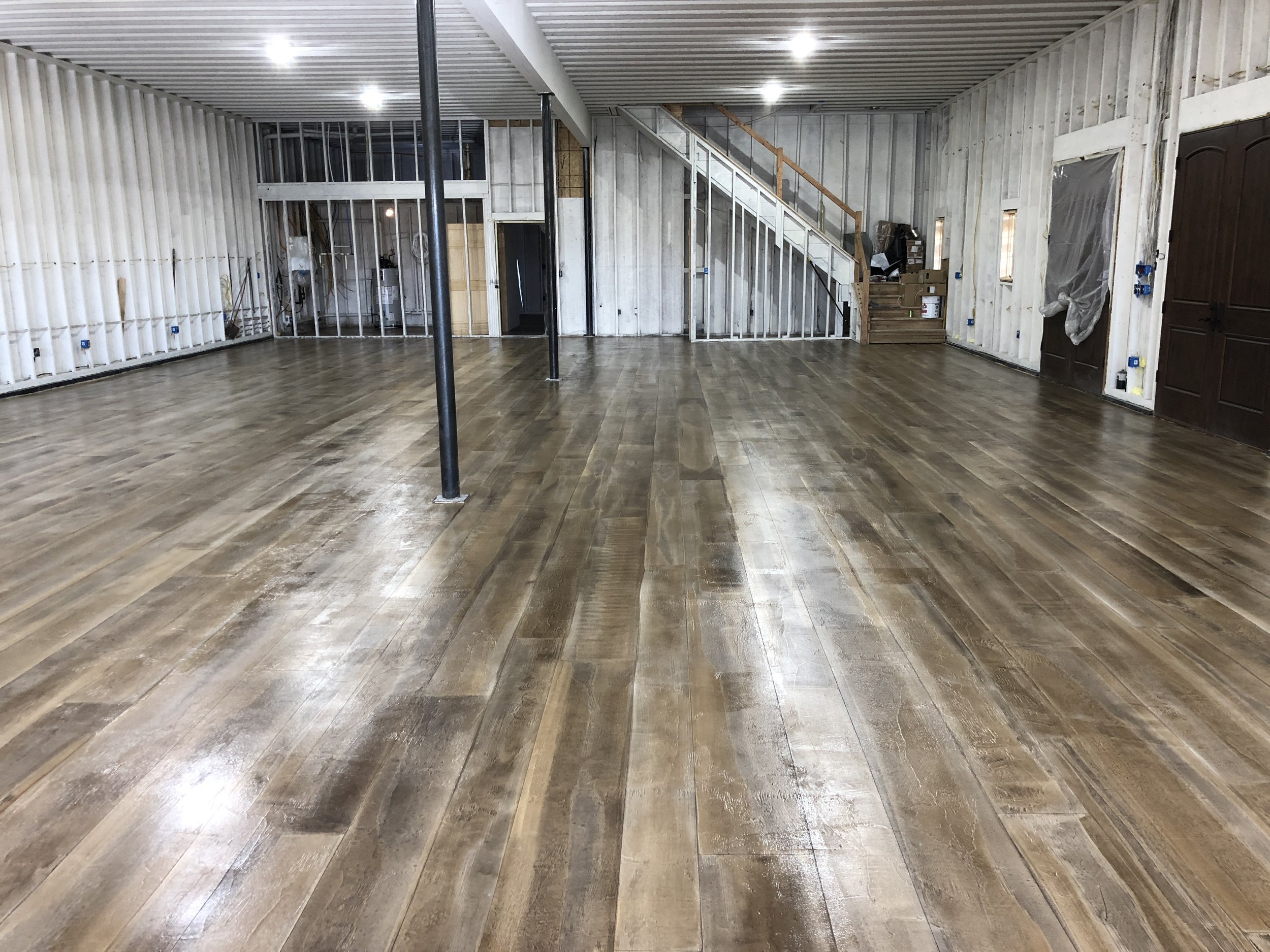 Interior of a large room under construction with newly installed wooden flooring, unfinished white panel walls, exposed electrical outlets, and a staircase leading to an upper level.