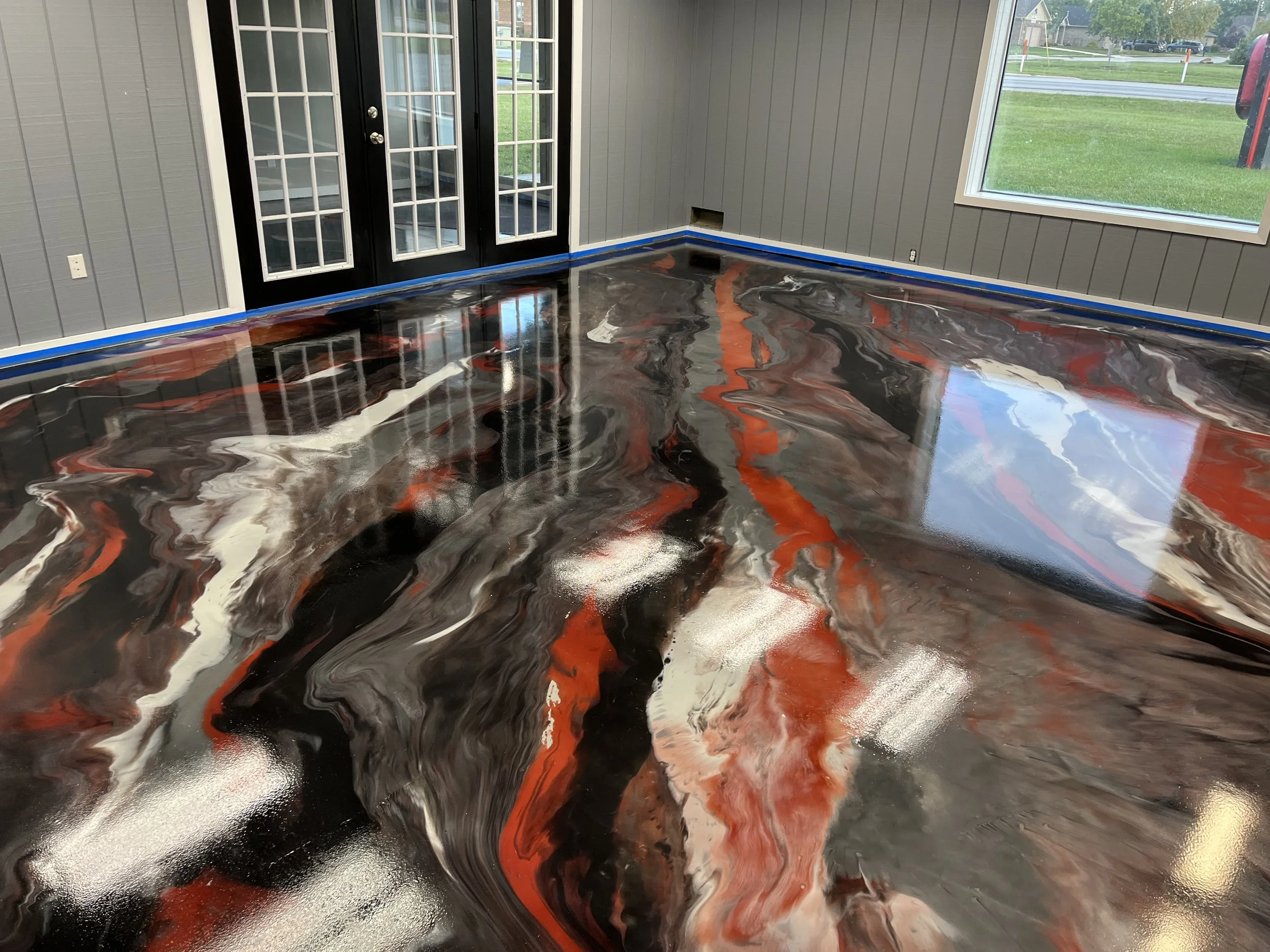 A room with a newly poured acrylic epoxy floor featuring swirls of black, gray, white, and red colors. The room has gray paneled walls, large windows, and black French doors leading outside.