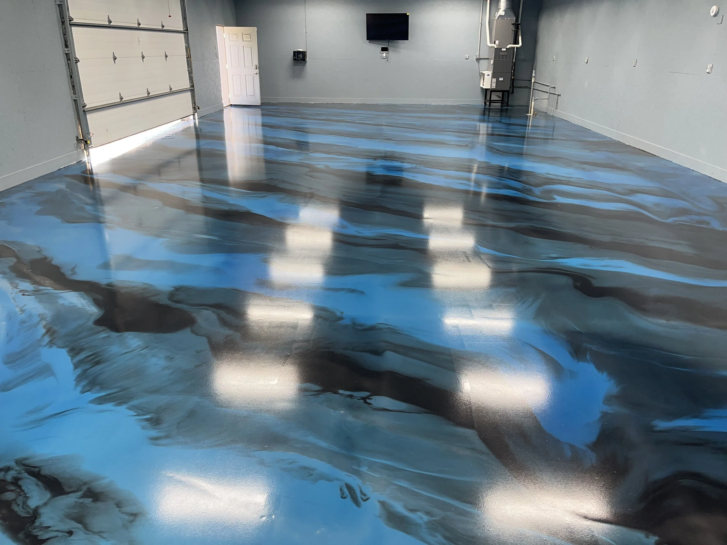 Empty garage with a refreshing blue and black marble-patterned epoxy floor, white garage doors on the left, a white door on the far wall, a wall-mounted TV, and electrical panels.