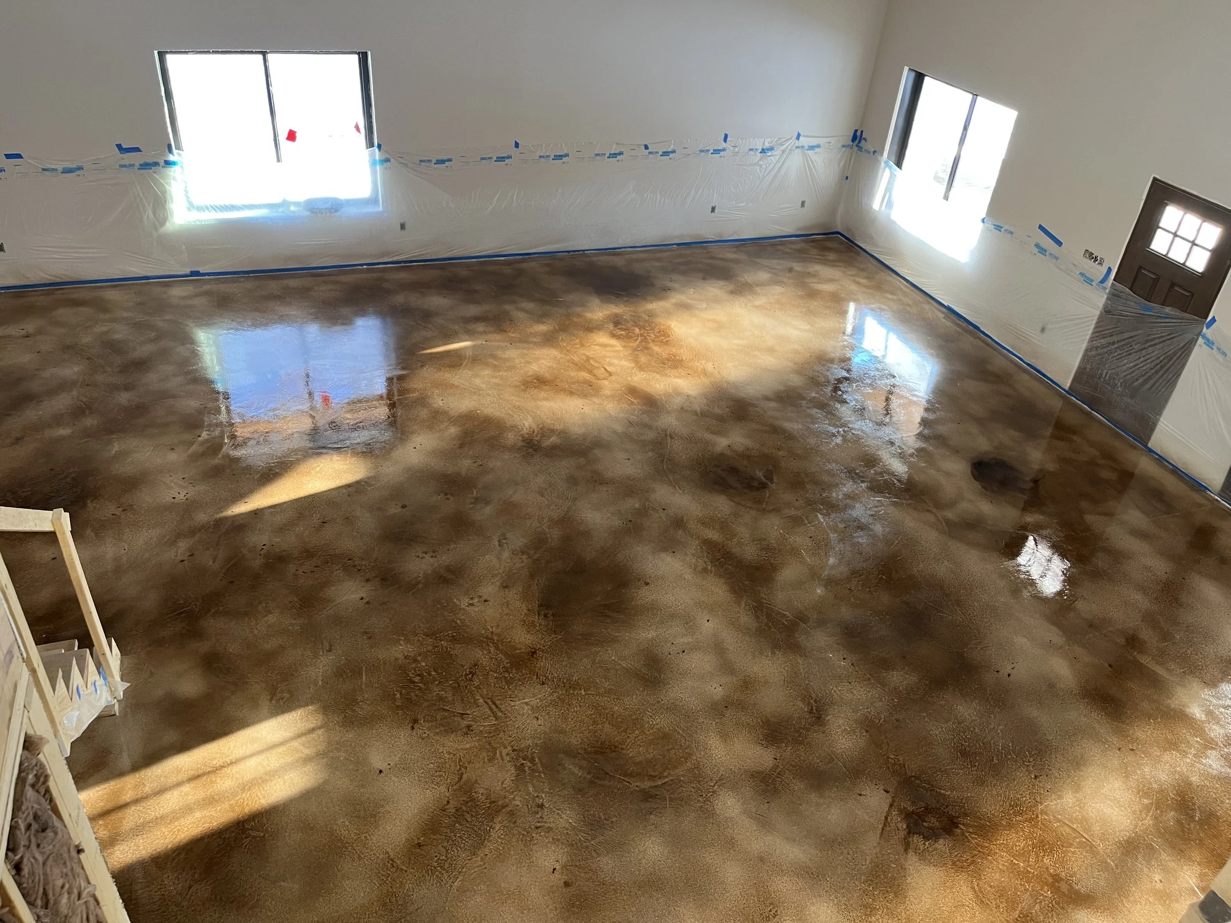Interior of a room under renovation with a stained and polished concrete floor, plastic sheeting on the walls, blue painter's tape, and windows letting in bright sunlight.