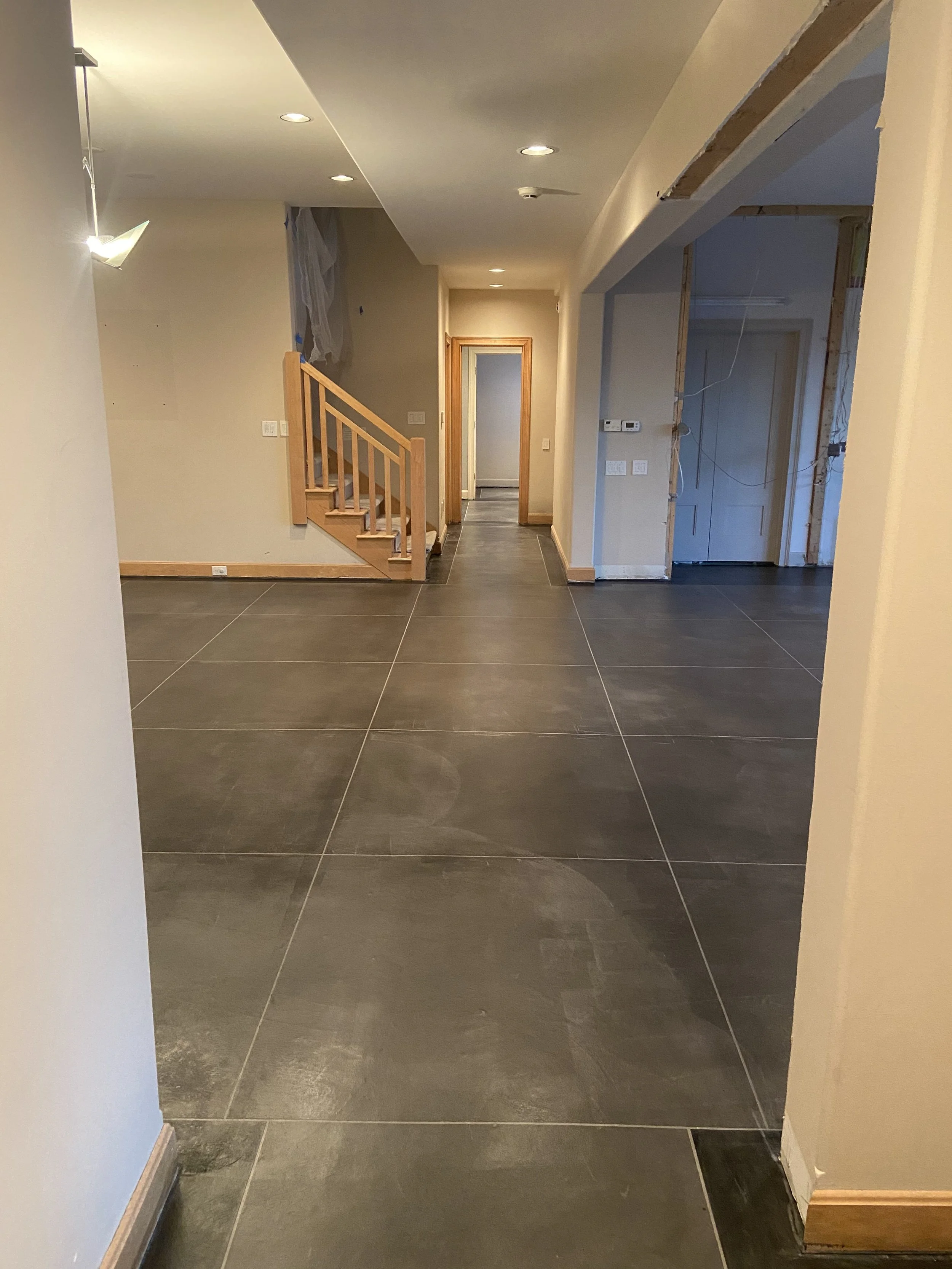 Interior of a house under renovation with dark tiled flooring, a staircase with wooden banister, and a hallway with a doorway at the end.