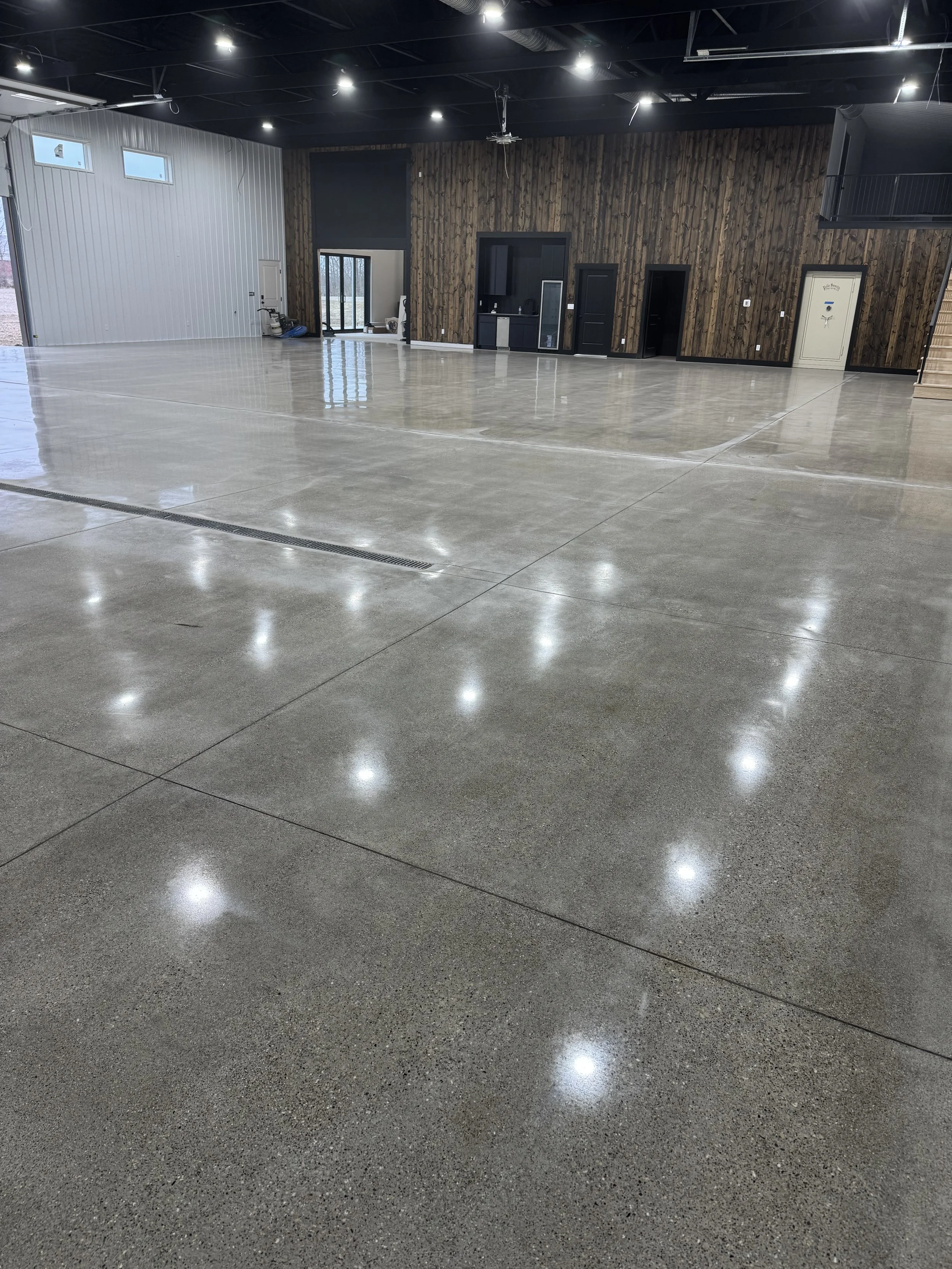 Empty indoor space with polished concrete floors, black-paneled walls, and a wooden accent wall. Ceiling has exposed black beams and lighting, with a staircase on the right side.