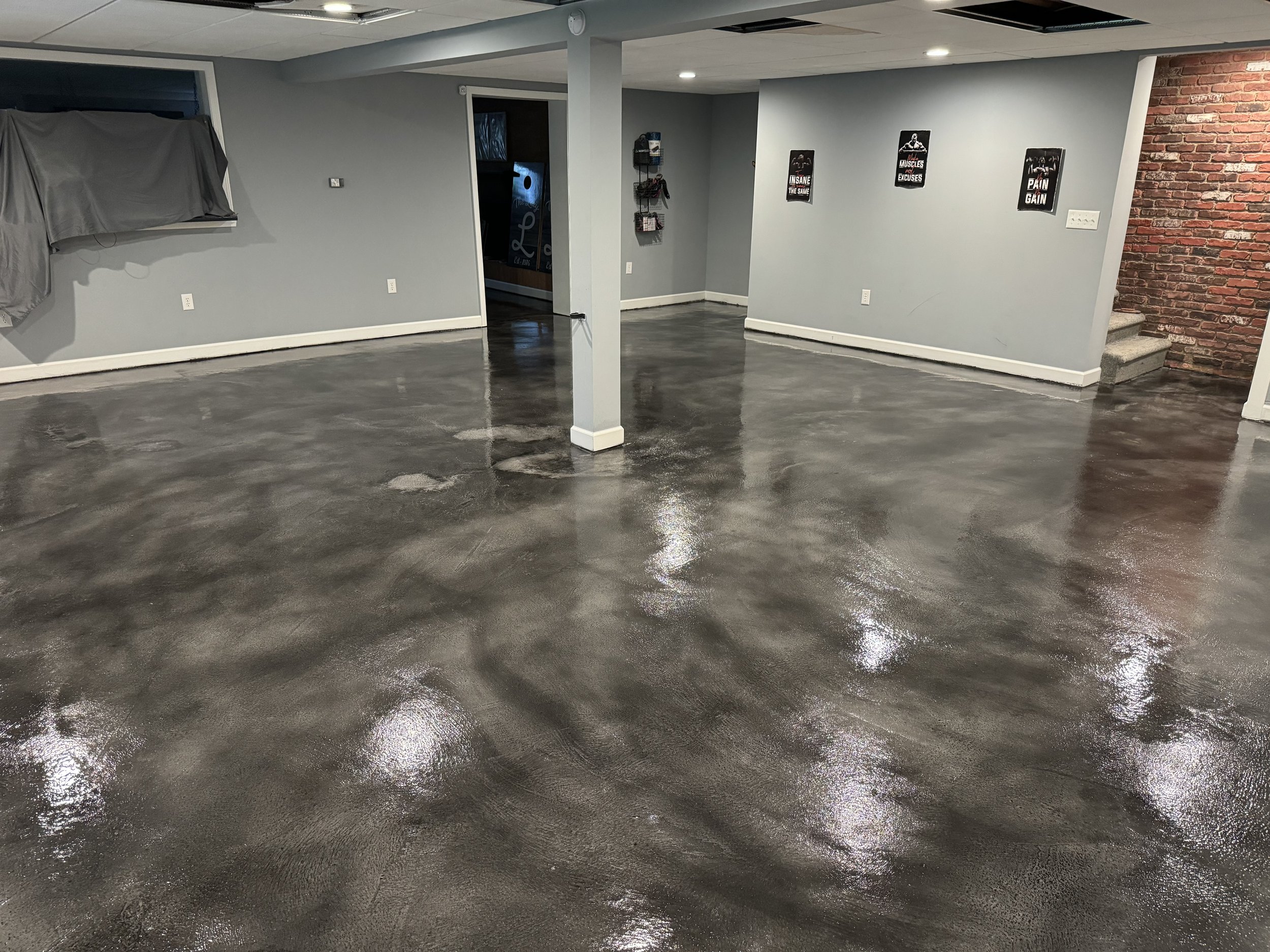Empty basement or garage with freshly polished concrete floor, gray walls, brick accent wall on the right, framed motivational posters on the walls, and a staircase leading upstairs.