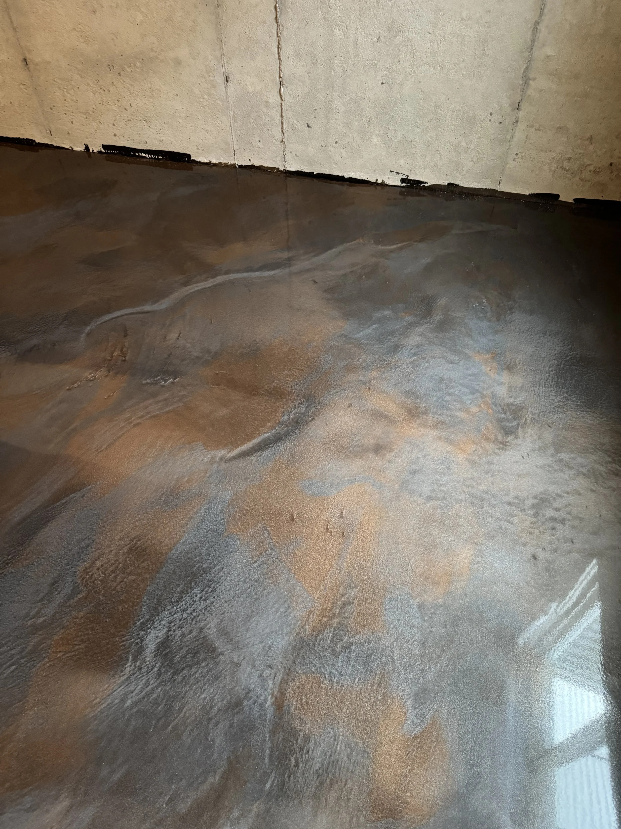 Close-up of a shiny, textured concrete floor with a miscellaneous stain and marks, against a concrete wall in a construction or renovation setting.