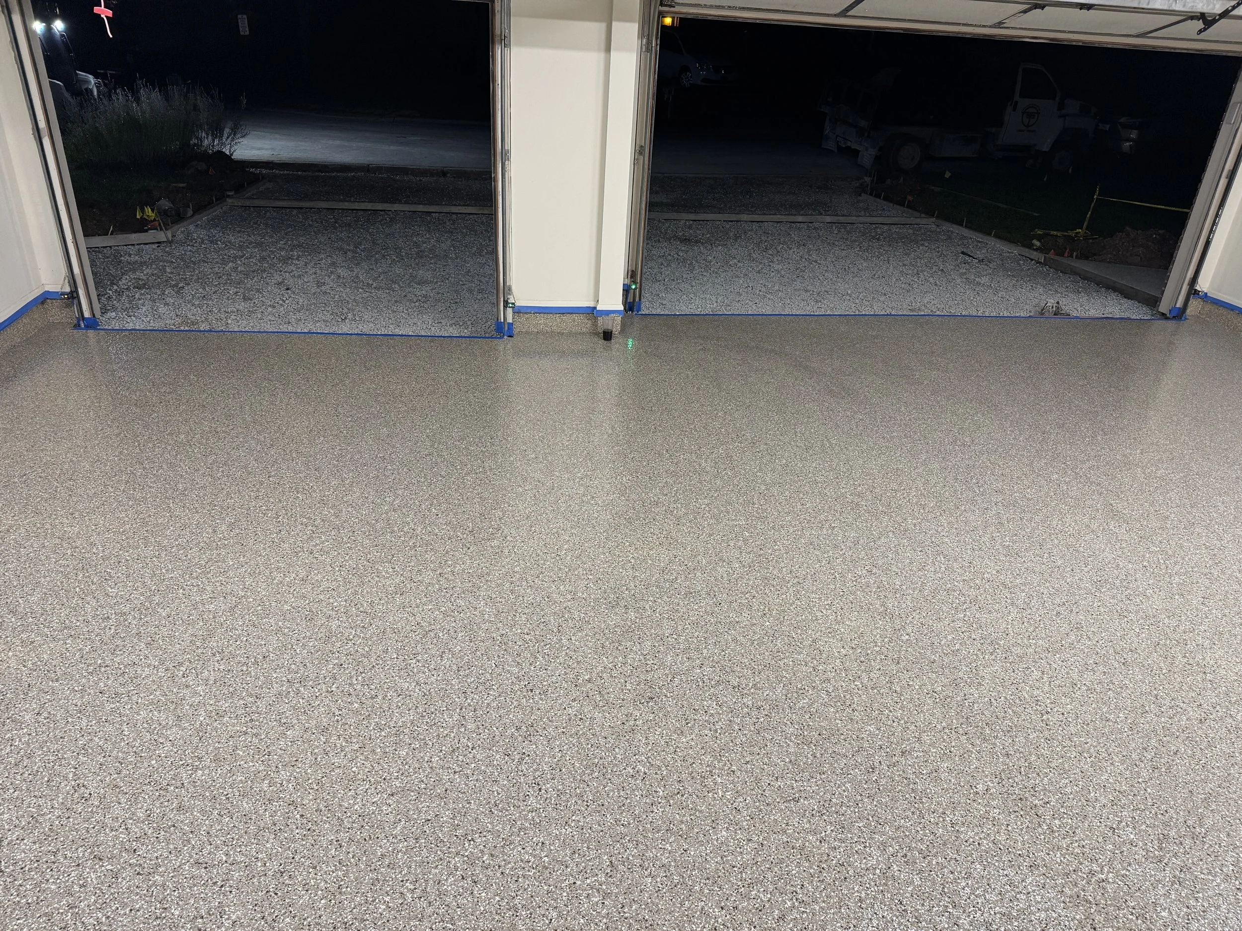 Interior of a garage with a newly finished speckled epoxy floor, with open garage doors revealing a dark outdoor area under construction.