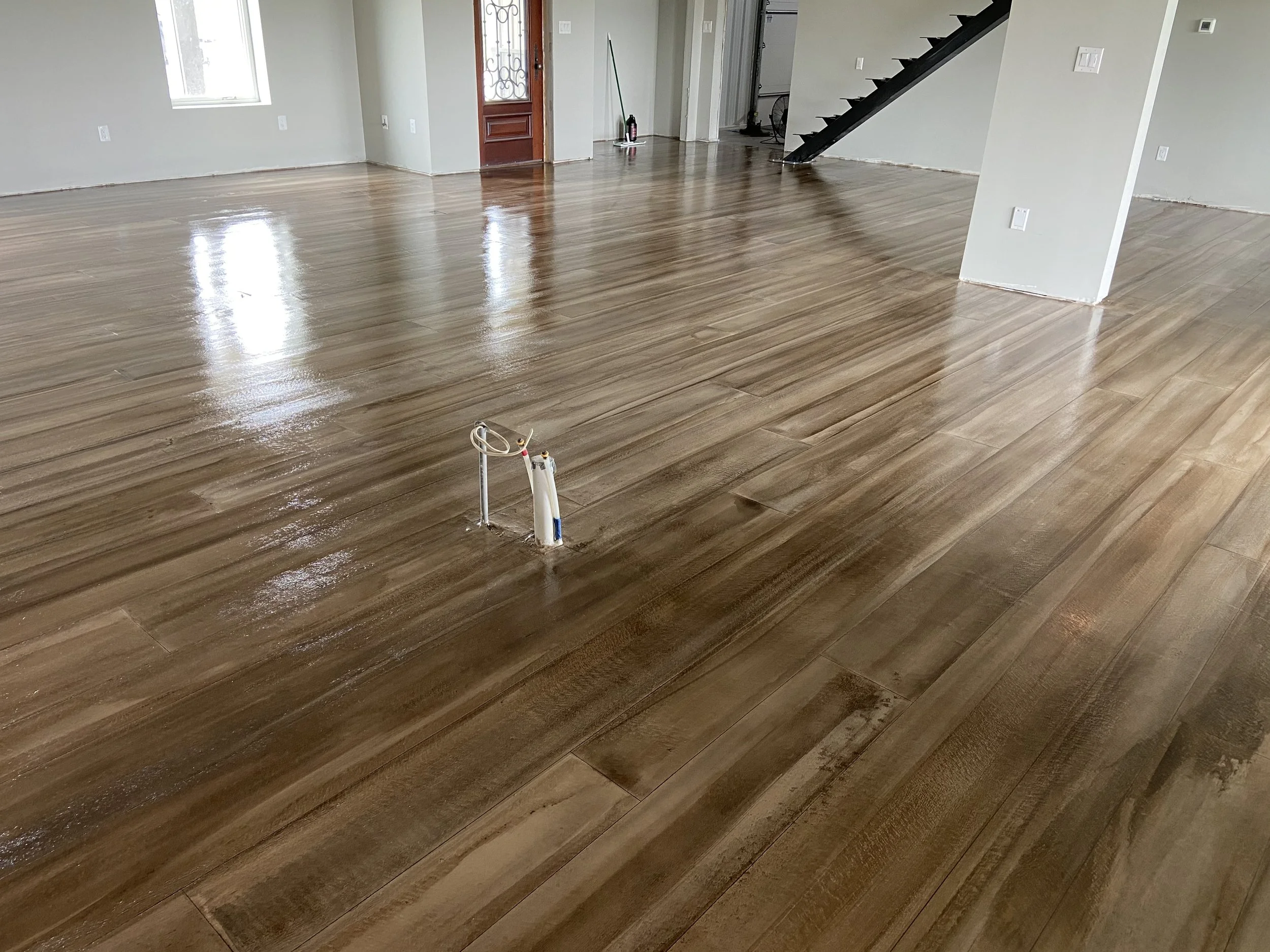 Empty living room with newly installed shiny wooden flooring, electrical wires protruding from the floor, and a staircase in the background.