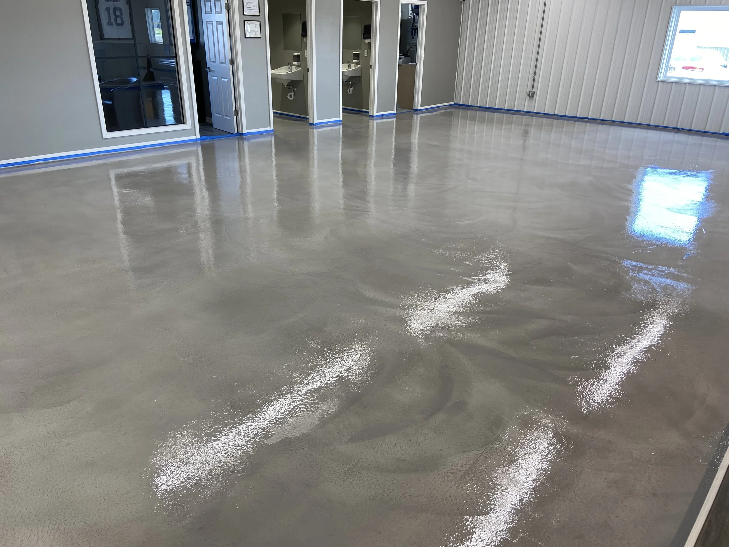 Empty room with a polished concrete floor, blue painter's tape along the baseboards, and a row of sinks in a small corridor.