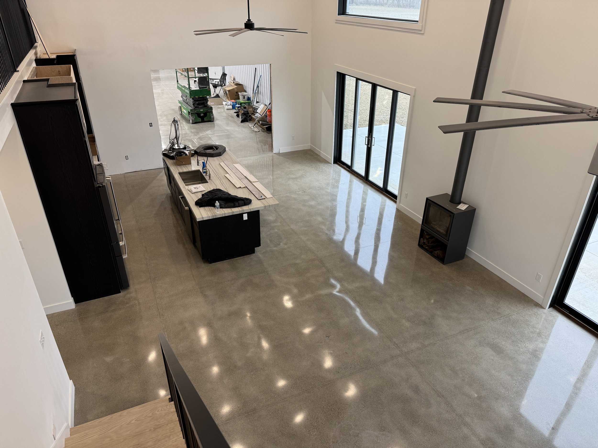 Empty living area with polished concrete floors, large sliding glass doors, a modern fireplace, and a kitchen island under construction, with tools and construction materials visible.