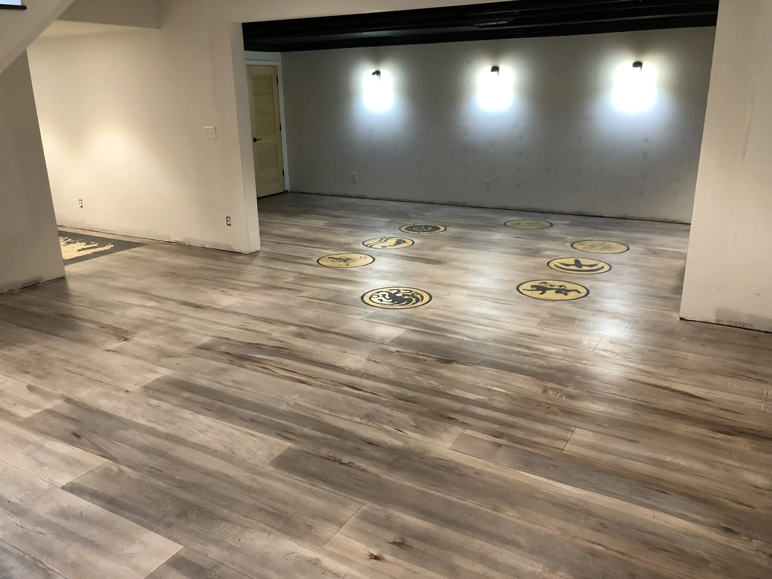 Empty hall with wood flooring and decorative floor medallions, white walls, and three wall-mounted lights on the far wall.