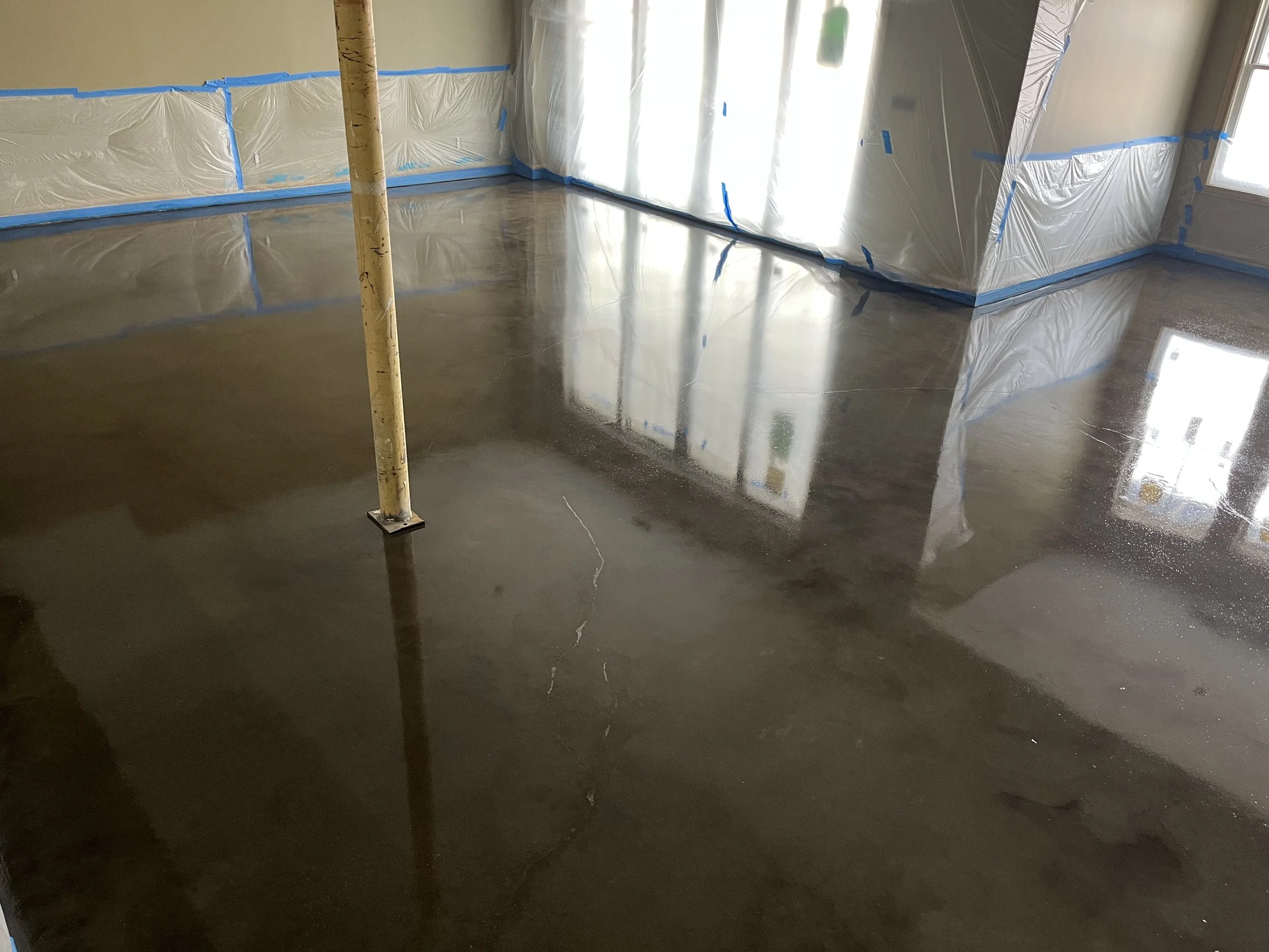 Interior room under construction with a freshly poured concrete floor that is shiny and wet, covered with plastic sheeting protected with blue painter's tape on the walls and around windows, and a support pole standing in the middle of the room.