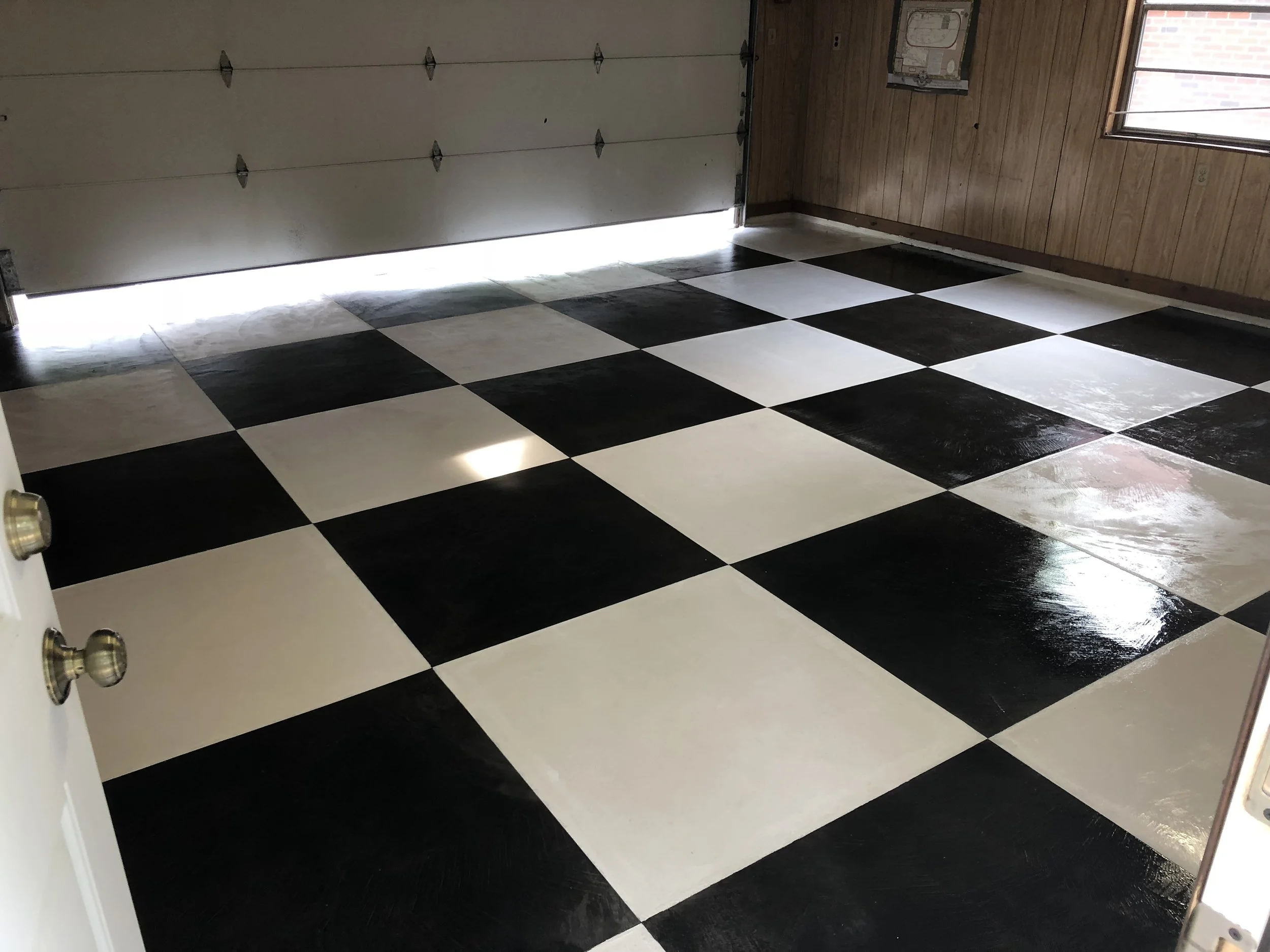 Clean garage with black and white checkered floor, white garage door, wood-paneled walls, window, and a small electrical panel.