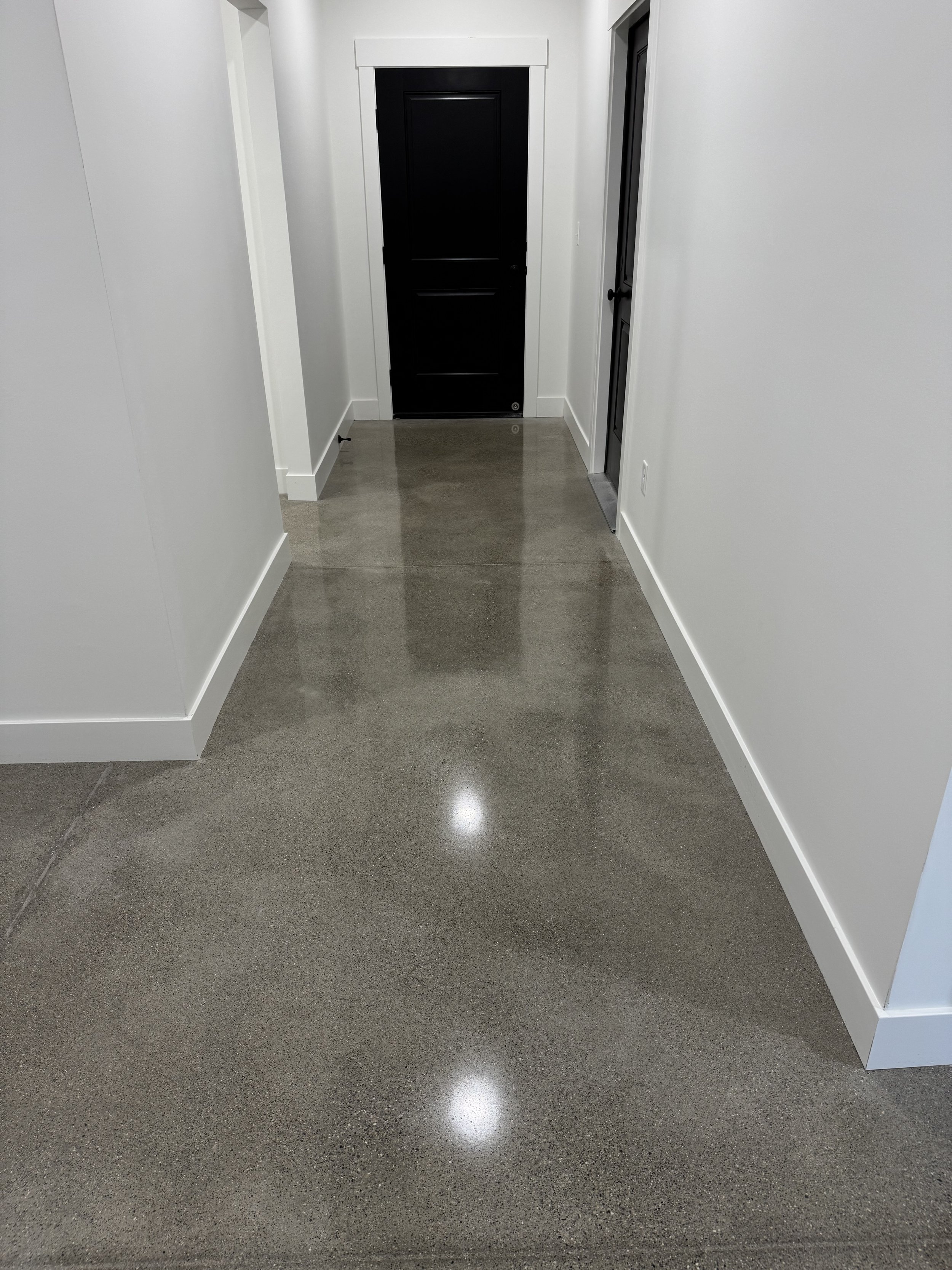 White hallway with polished concrete floor leading to a black door at the end and a glass-paneled door on the right.