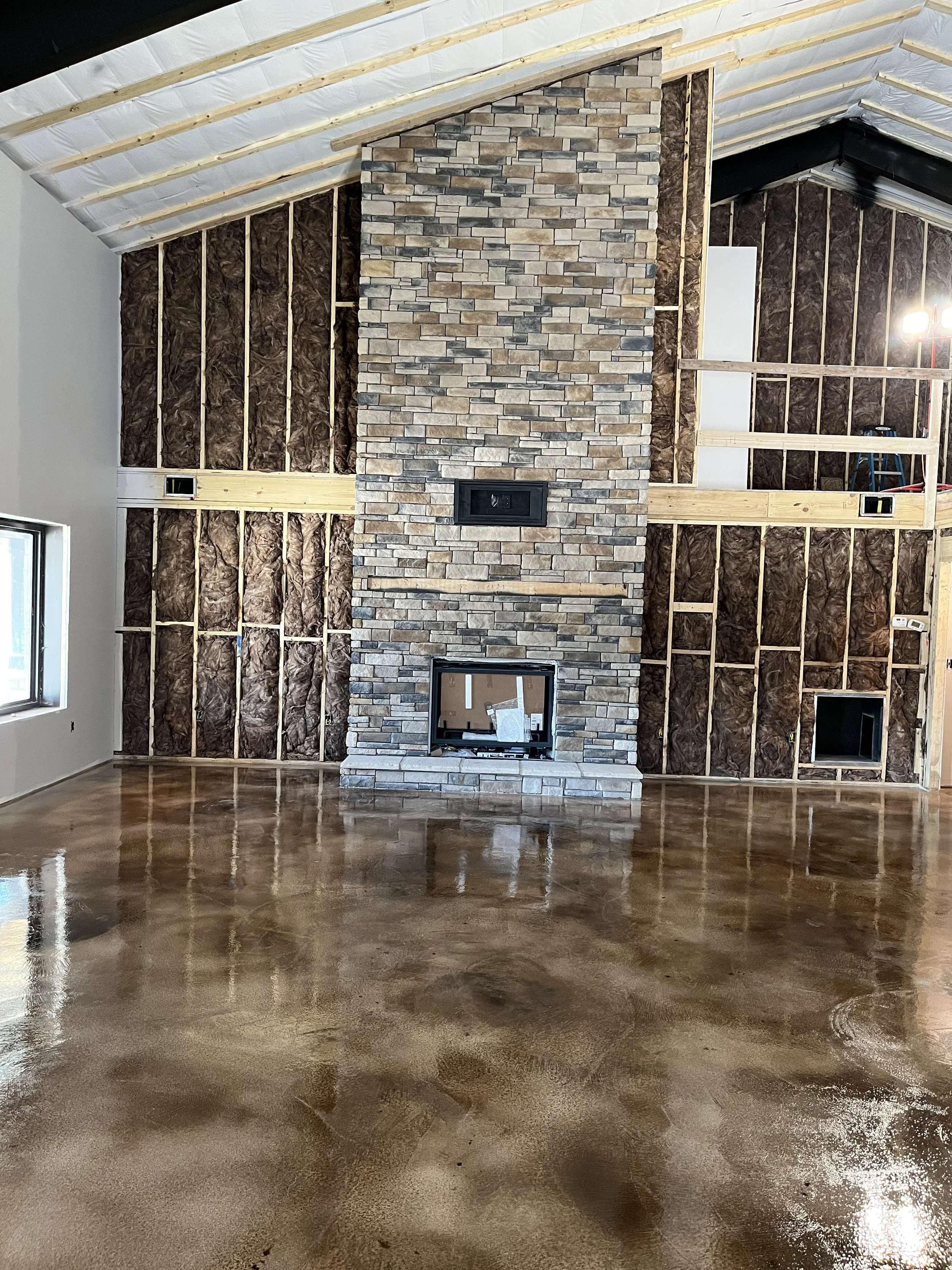 Living room under construction with exposed wall framing, a central stone fireplace, and polished concrete floor.