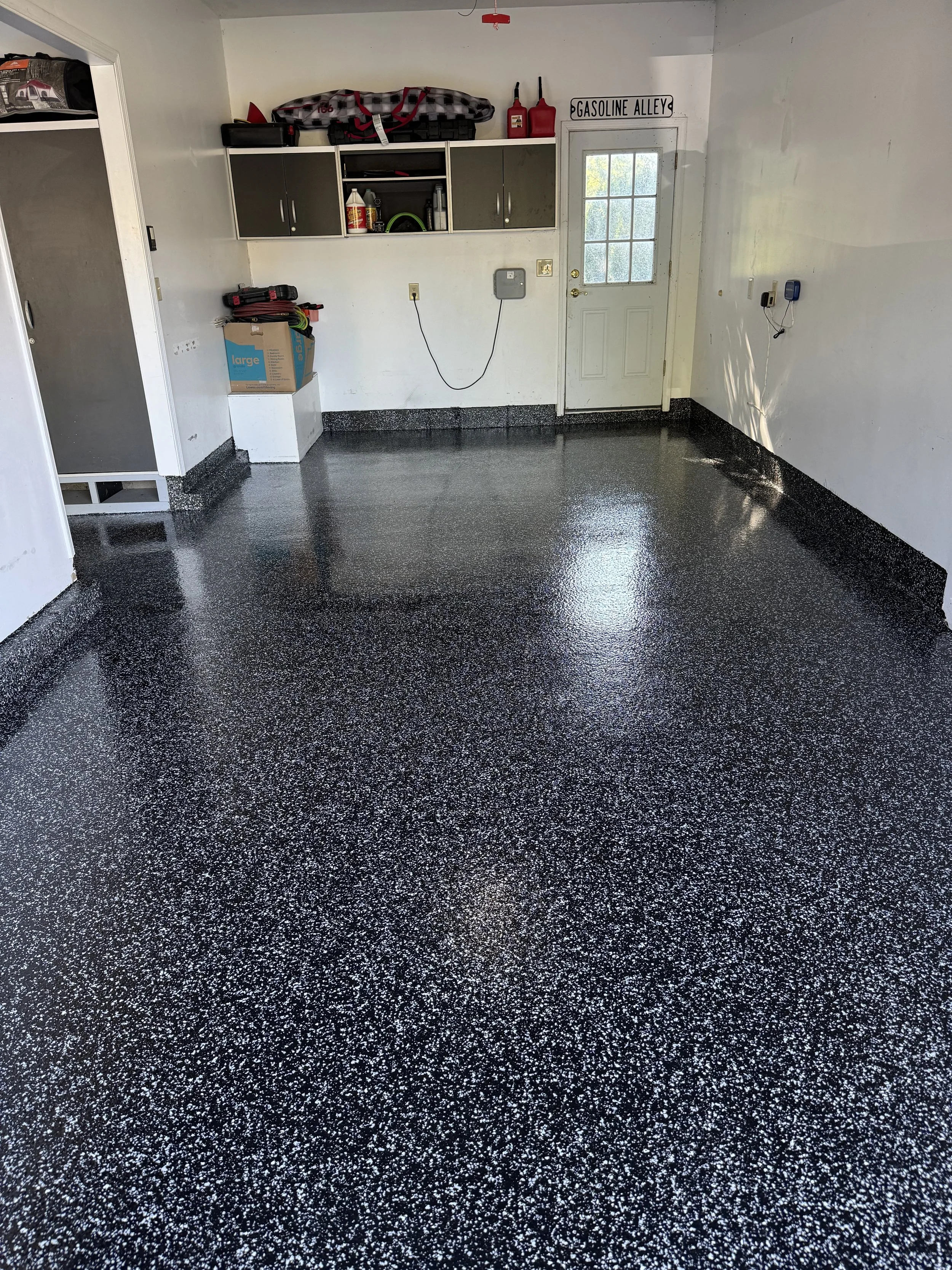 Empty garage with black and white speckled epoxy coated floor, white walls, and a storage area with cabinets, shelves, and various items.