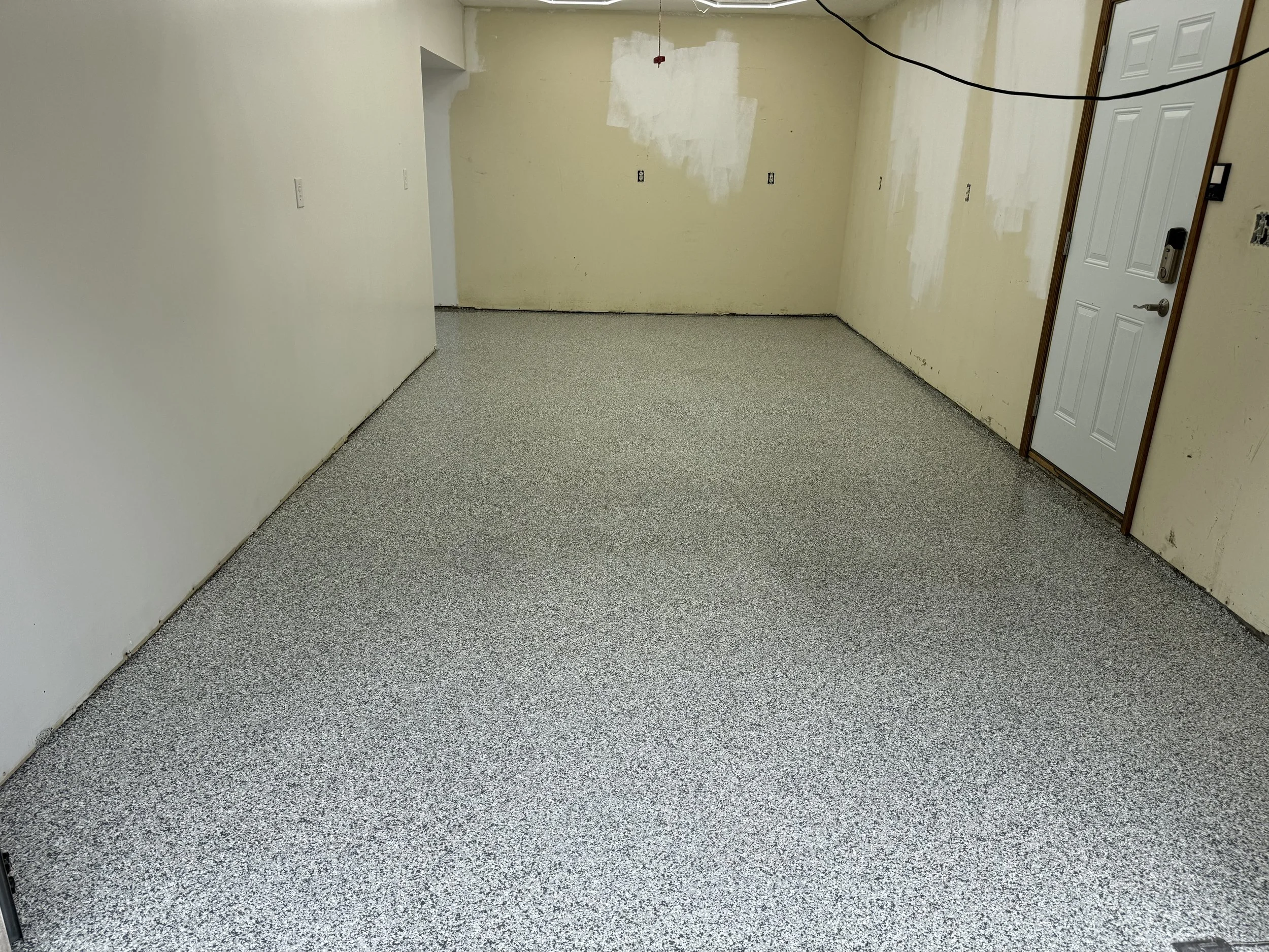 Empty room with new speckled gray and white flooring, unfinished walls with patches of primer, a white door with a lock, and exposed wiring hanging from the ceiling.
