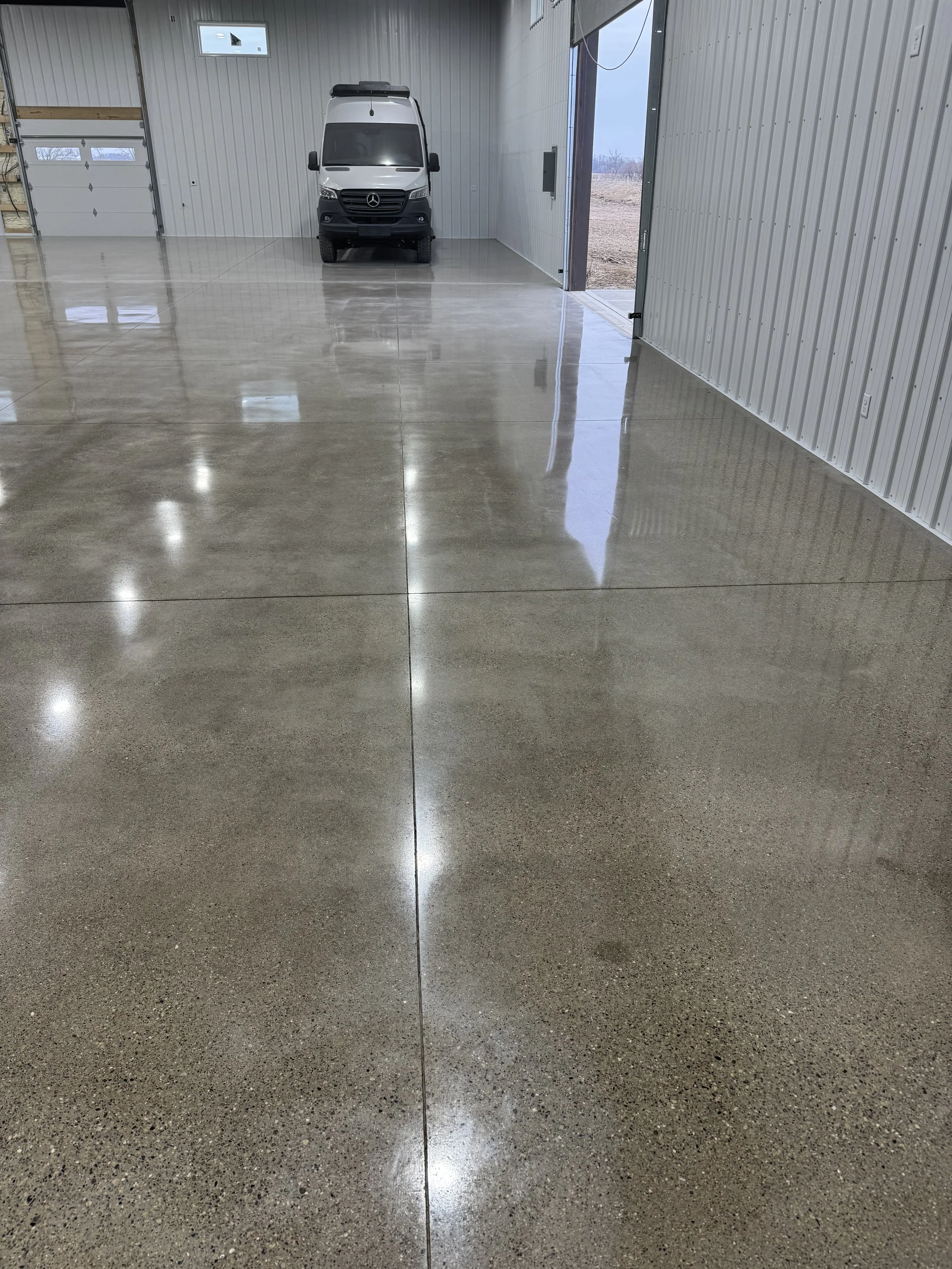 Empty garage with a polished concrete floor, white metal walls, a Mercedes-Benz van parked near the rear wall, and an open garage door showing an outside view of a dirt field.