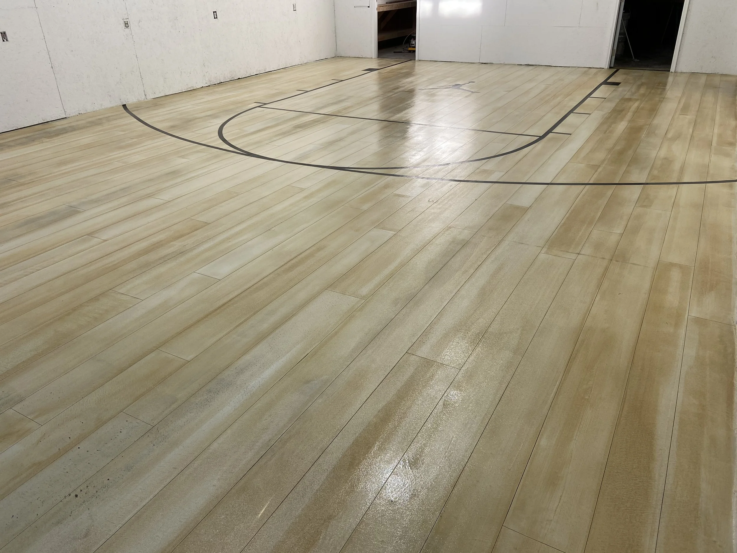 Indoor basketball court with light-colored wood flooring and black court markings, with unfinished walls and visible electrical outlets.