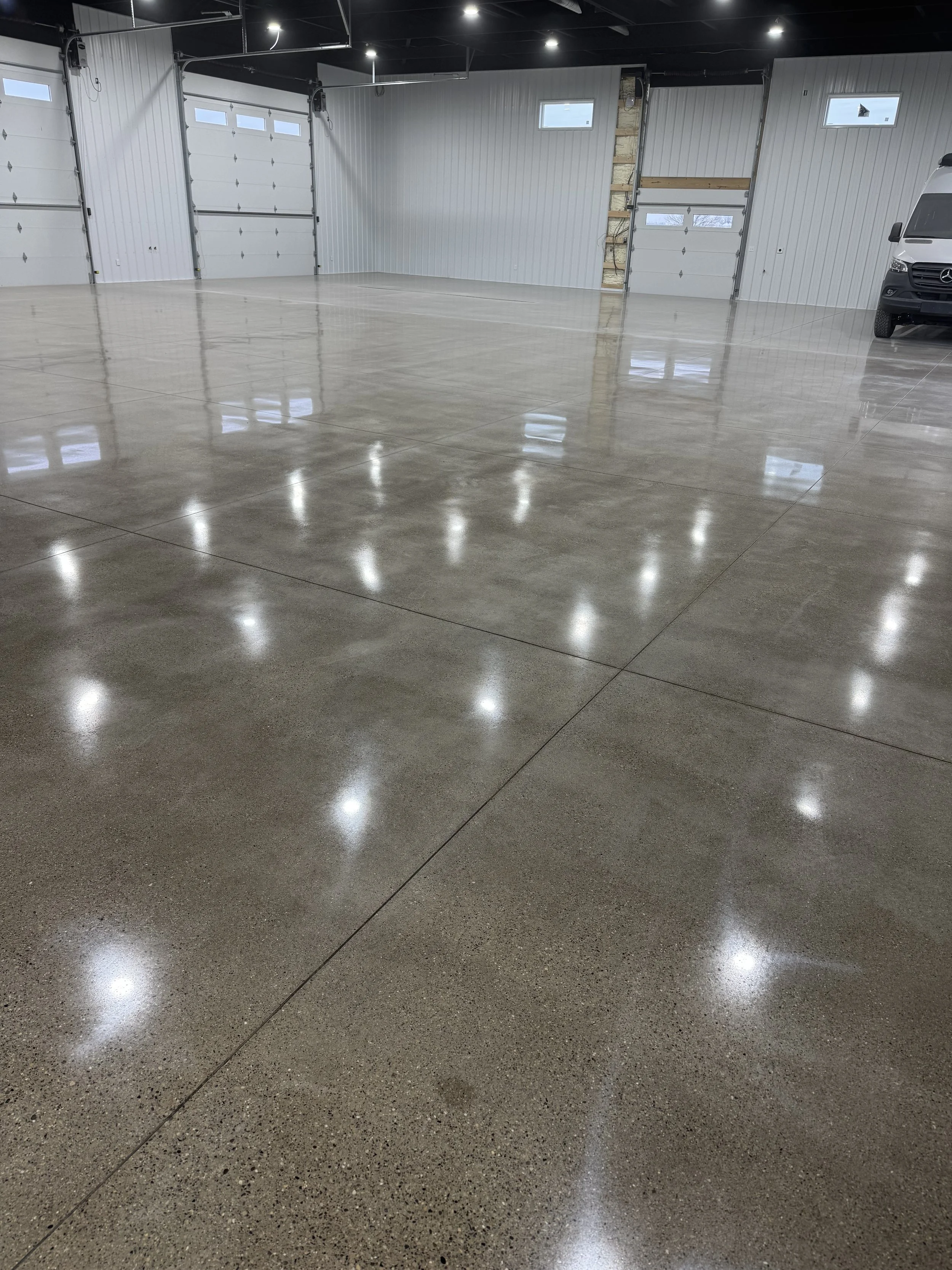 Empty warehouse with polished concrete floors, white metal walls, and garage doors, with a black van partially visible on the right side.