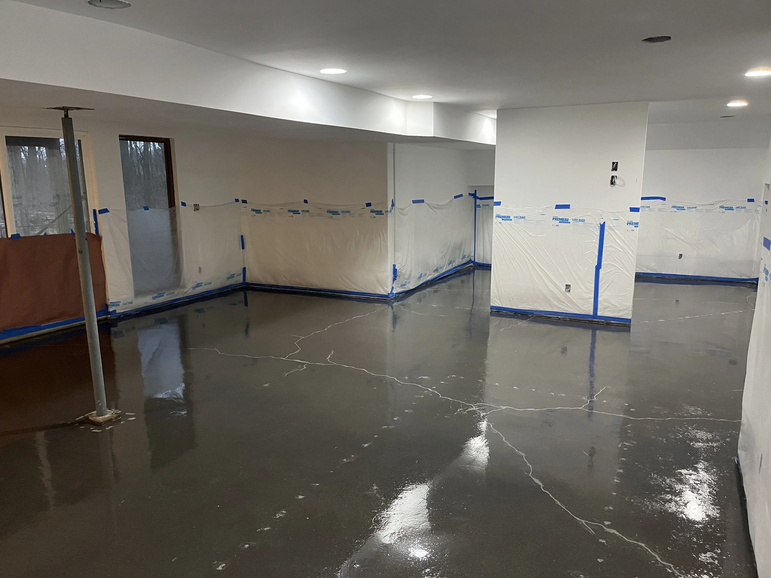 Interior of a room under renovation with freshly poured and polished concrete floor marked with cracks, partially covered windows, and walls covered with protective plastic sheeting.