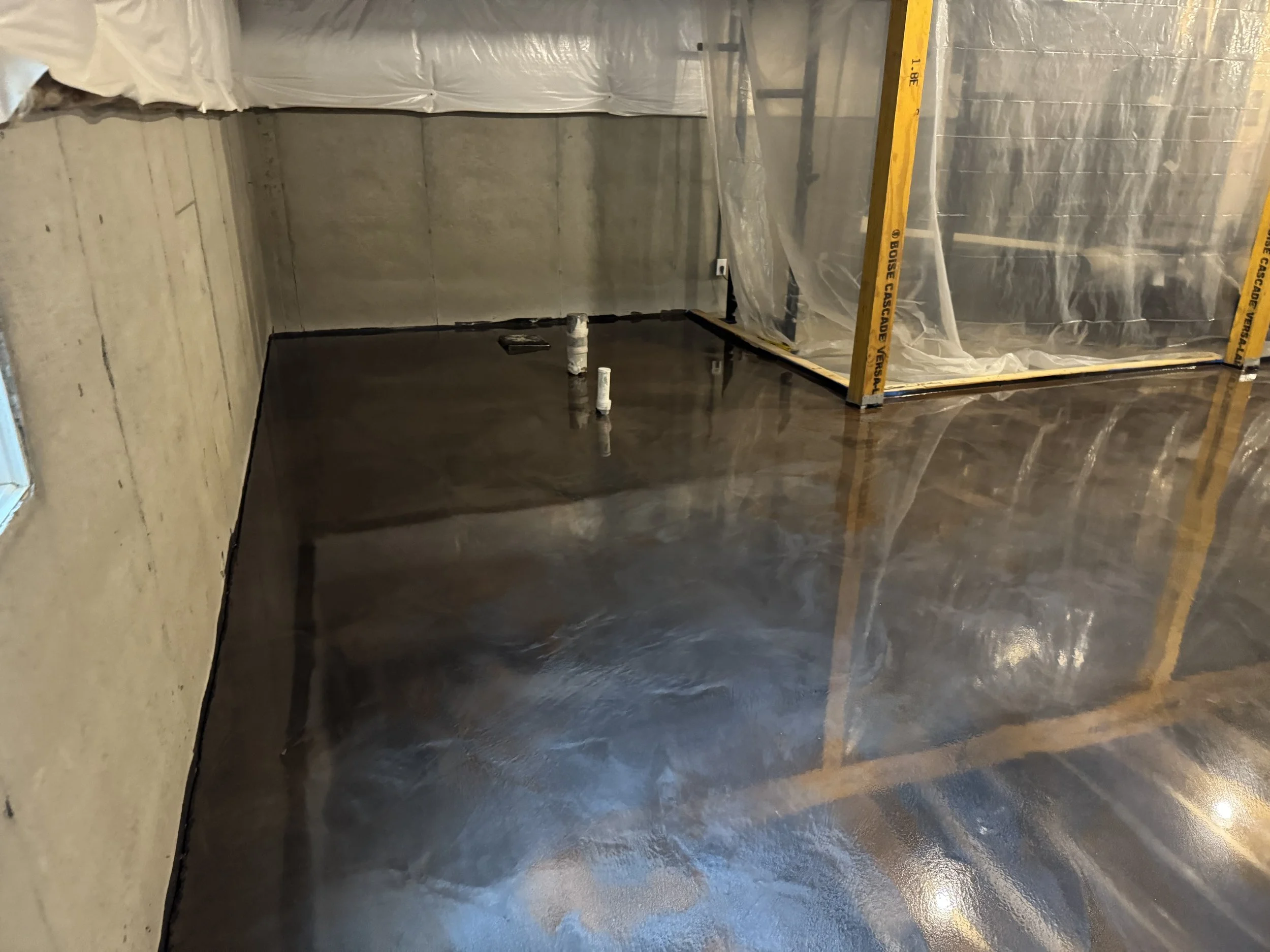 Under construction room with a newly poured concrete floor that is wet and reflective, with plumbing pipes and construction frames visible.