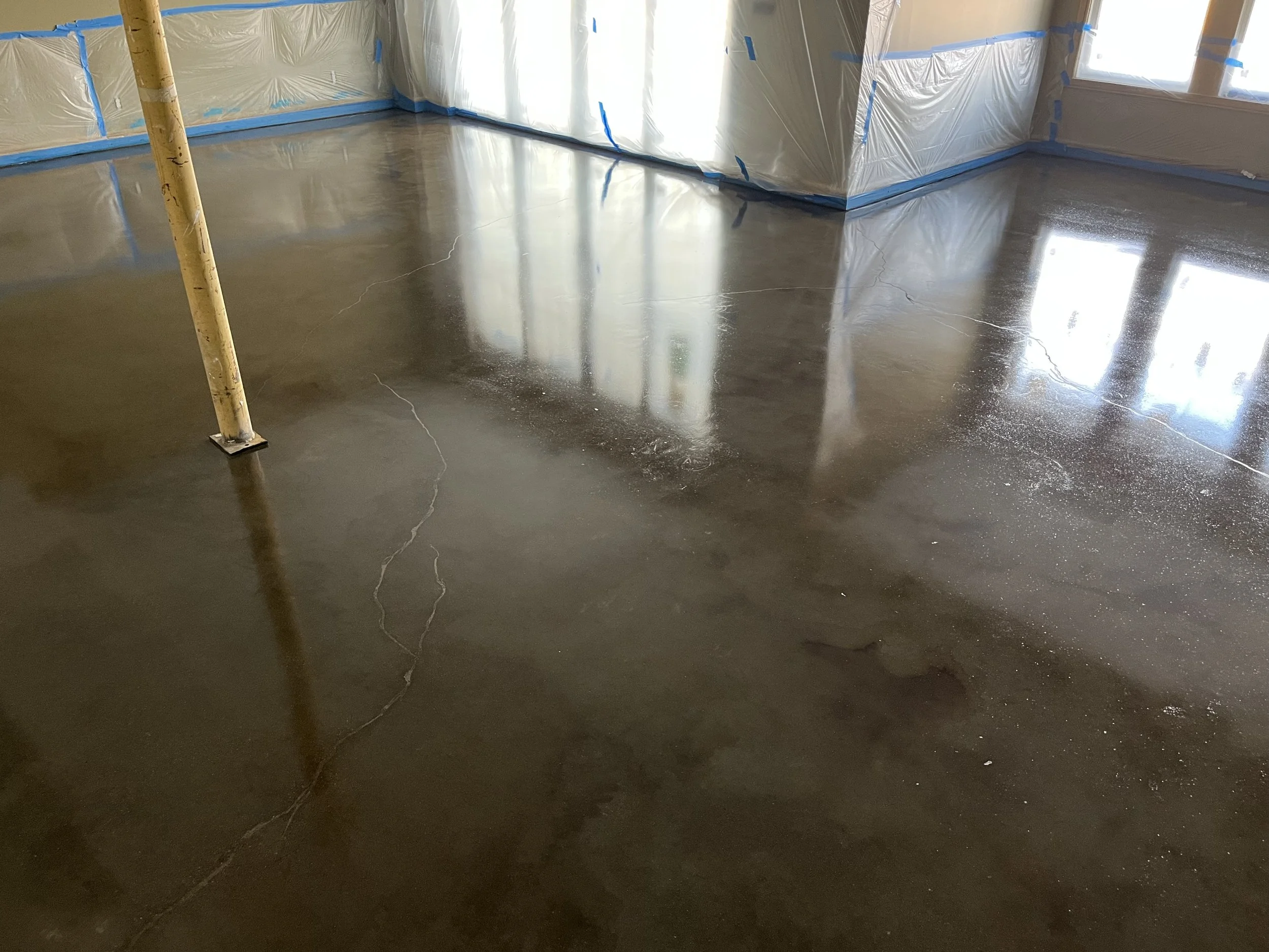 Interior of a room under construction with a freshly poured concrete floor that has visible cracks, plastic sheeting on the walls, and windows reflecting sunlight.