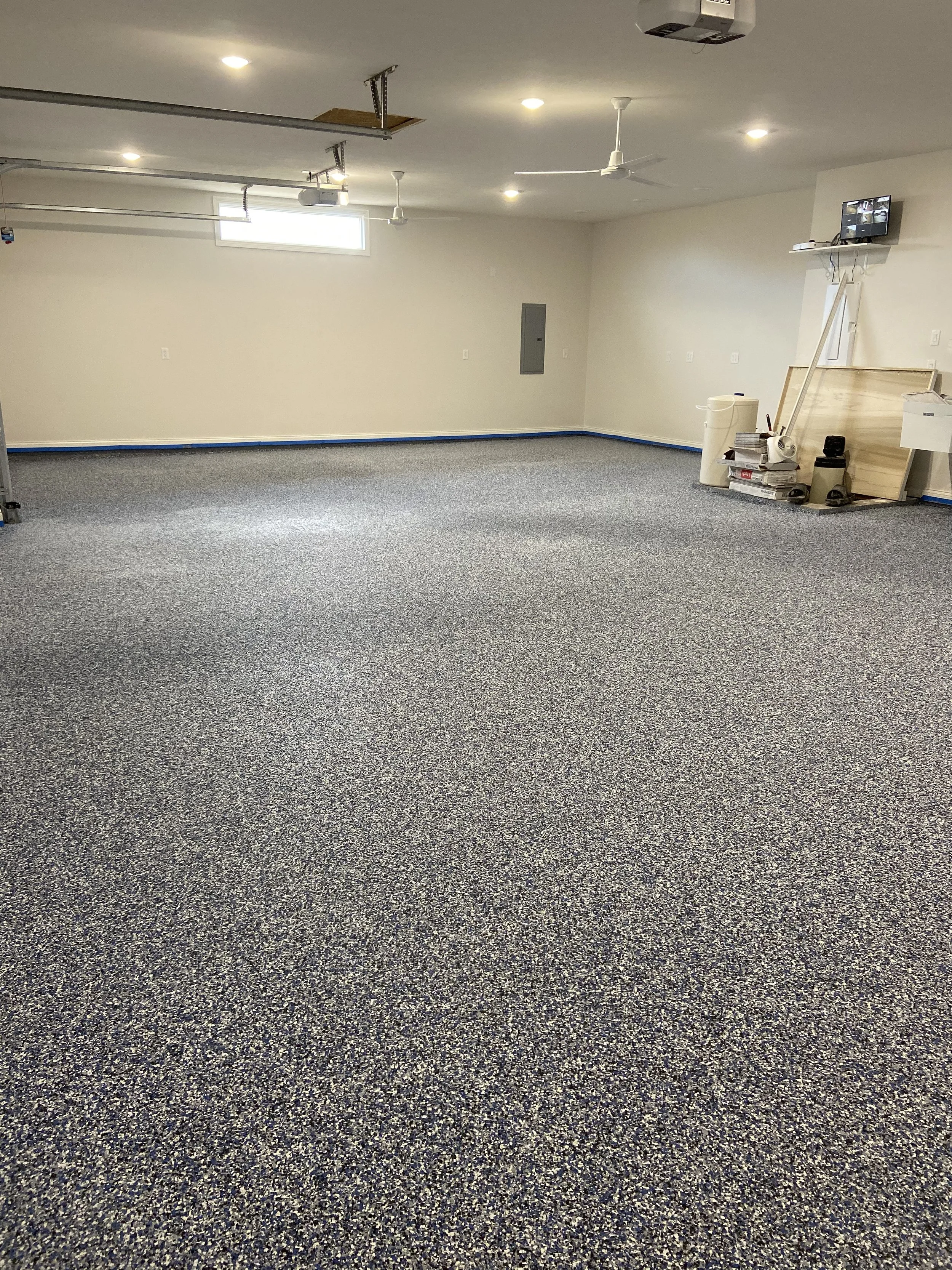 Empty garage or workshop space with speckled gray carpet flooring, white walls, recessed lighting, a small high window, and various tools and materials stacked in one corner.