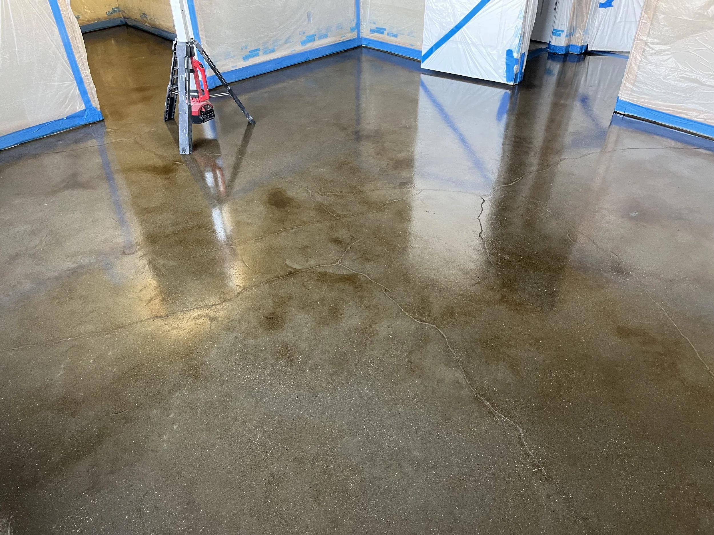 Polished concrete floor with cracks in a room under construction, partially covered walls with blue painter's tape, and a ladder with a red level in the background.