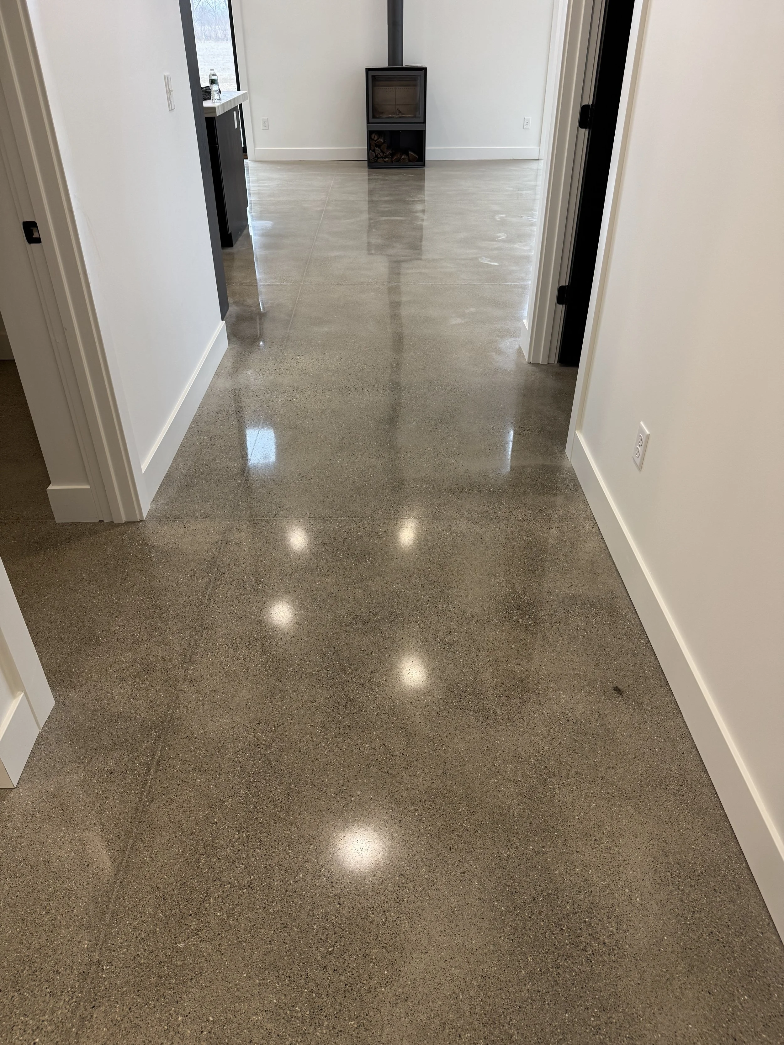 Interior view of a modern indoor space with polished concrete floors, white walls, and a small black fireplace with wood logs underneath. There is a window in the background letting in natural light.