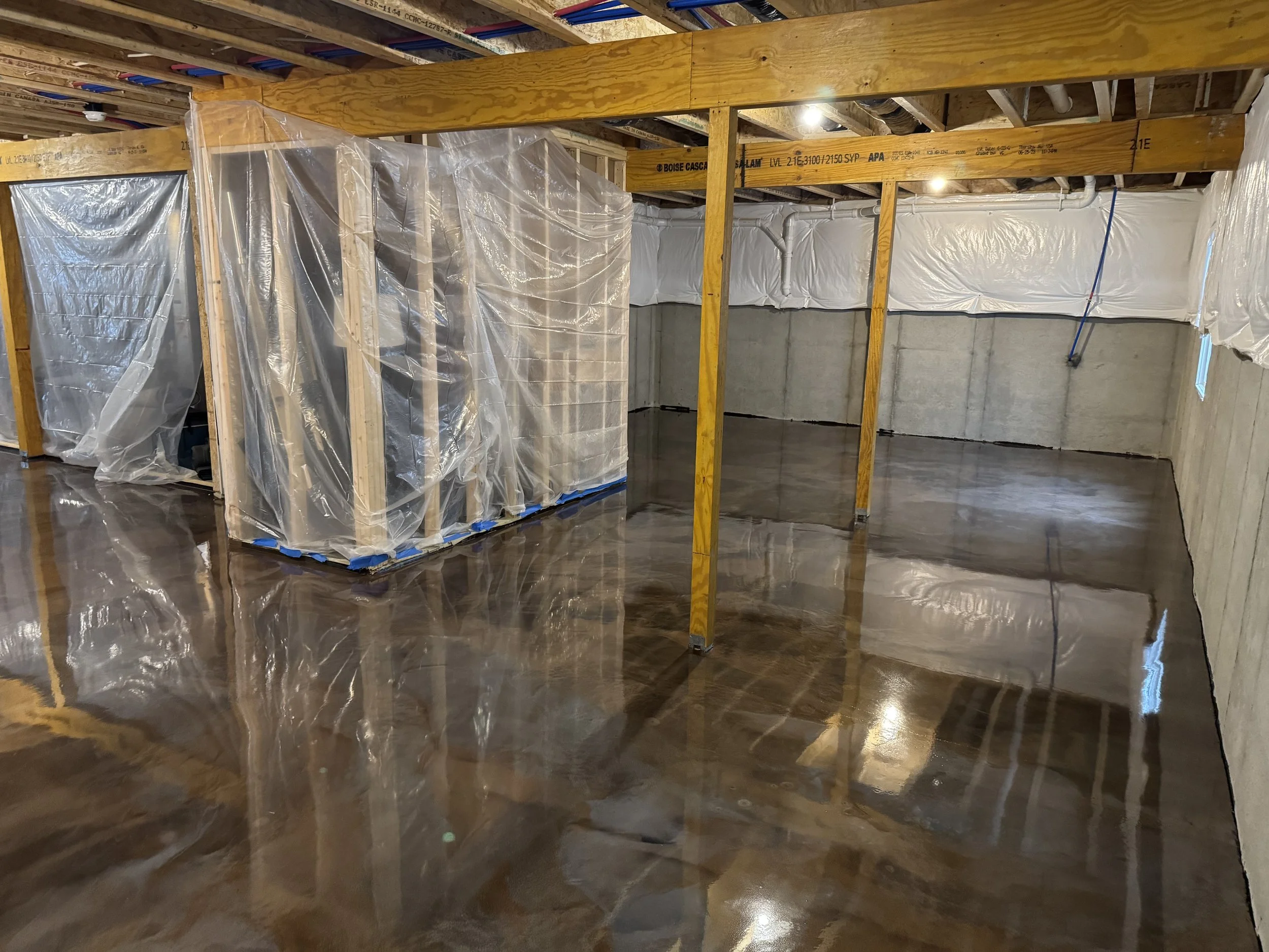 Interior view of a basement with a freshly poured concrete floor, exposed wooden framing, and insulated walls covered with white plastic sheeting.