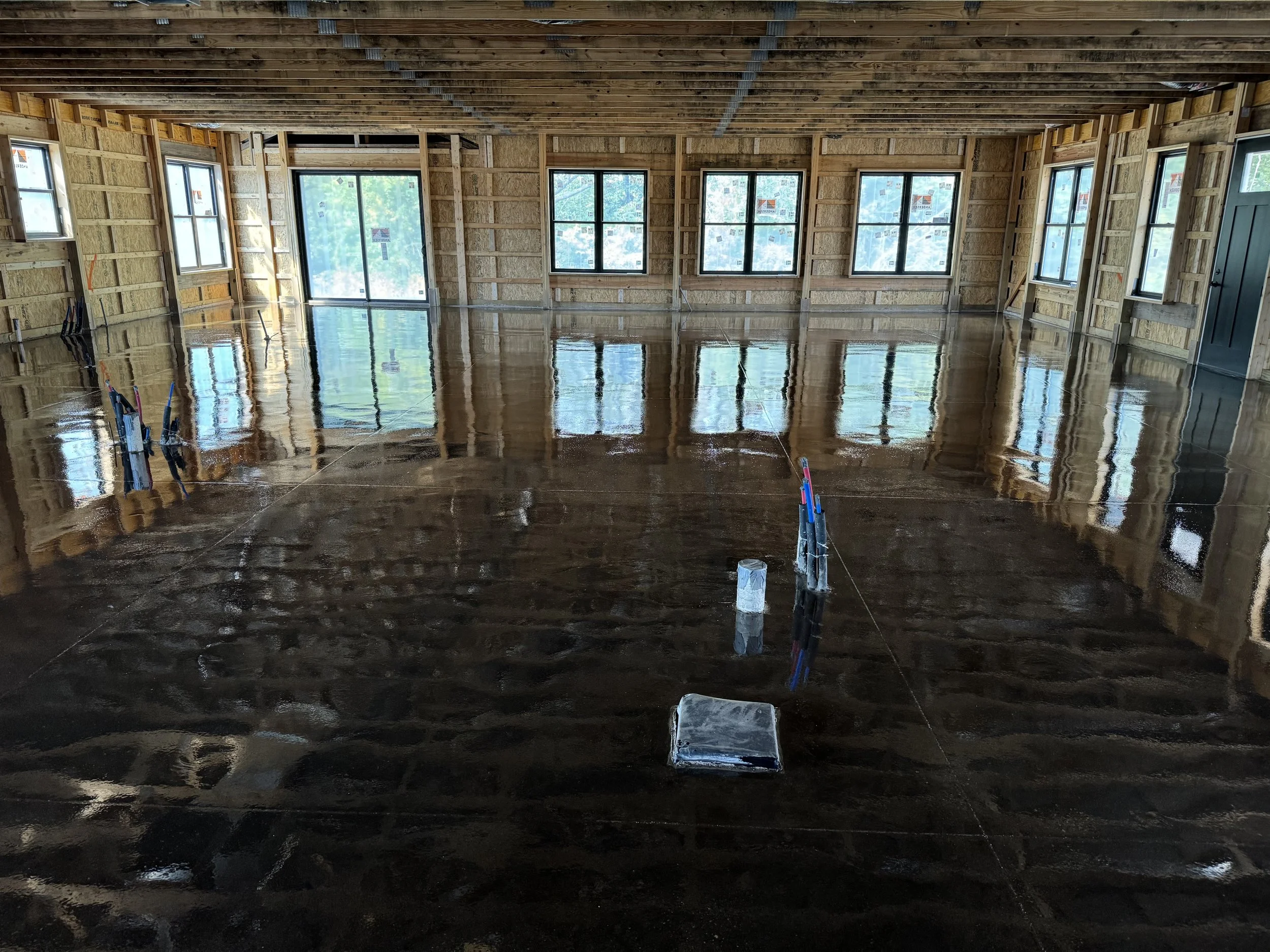 Interior of a building with exposed wooden framing and large windows, featuring a freshly poured and still wet dark-colored concrete floor that reflects the windows.