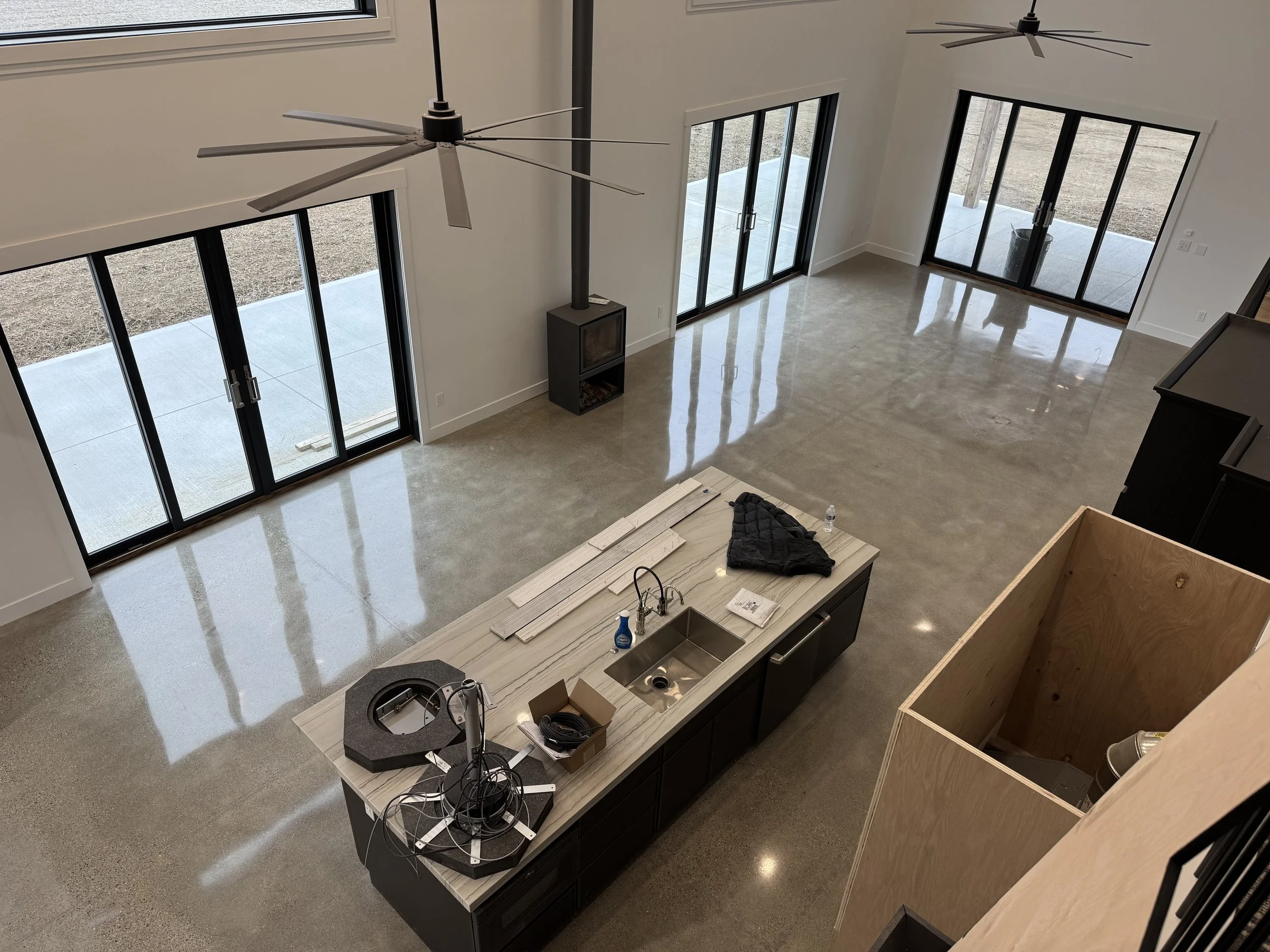 Empty living area with large sliding glass doors, a kitchen island with a sink, items on the counter, and a wood-burning stove in corner, possibly under construction.