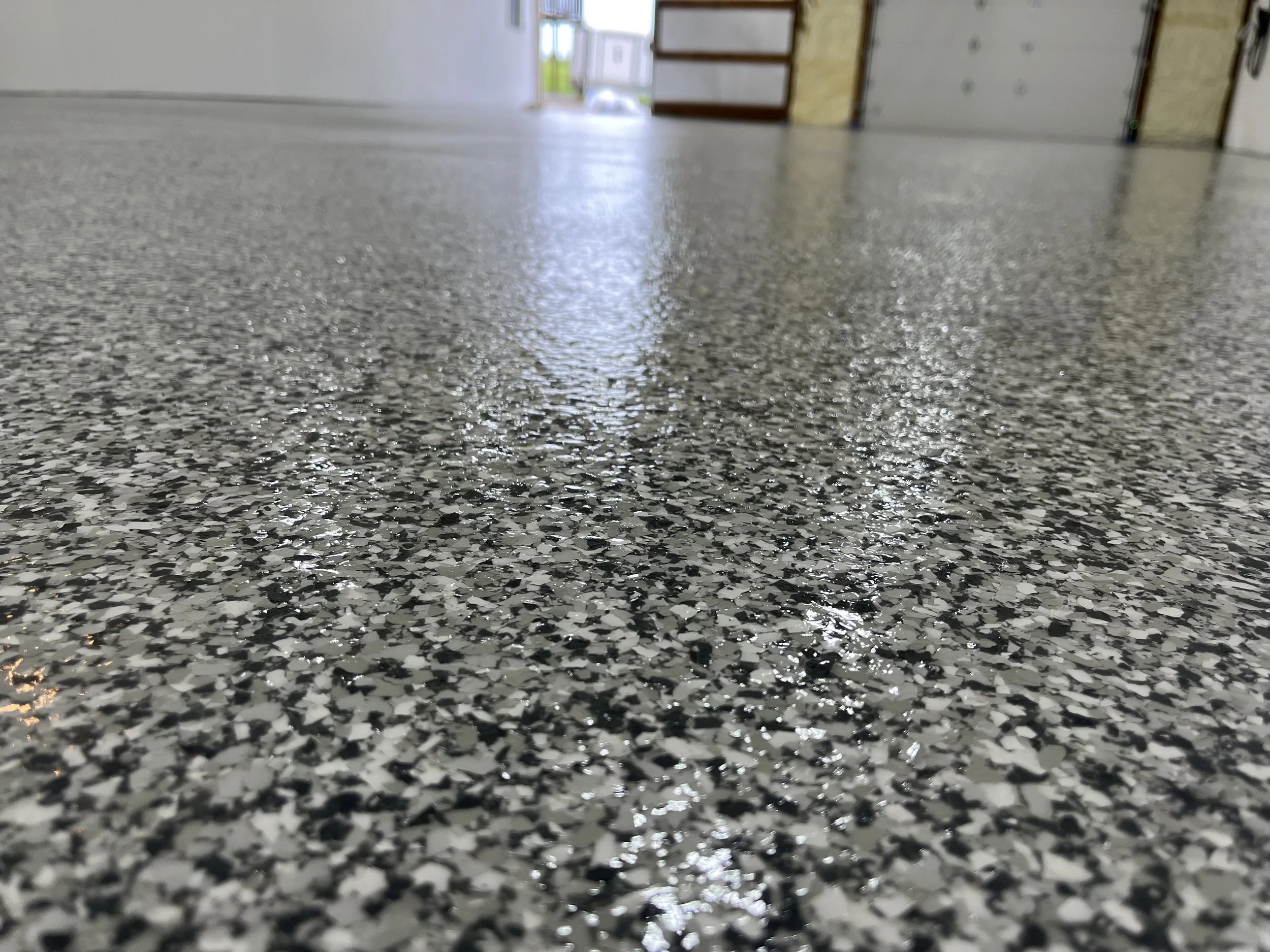 Close-up of a speckled gray floor with a reflection of light, showing texture and pattern. In the background, there are doors and a wall, with some natural light coming in.