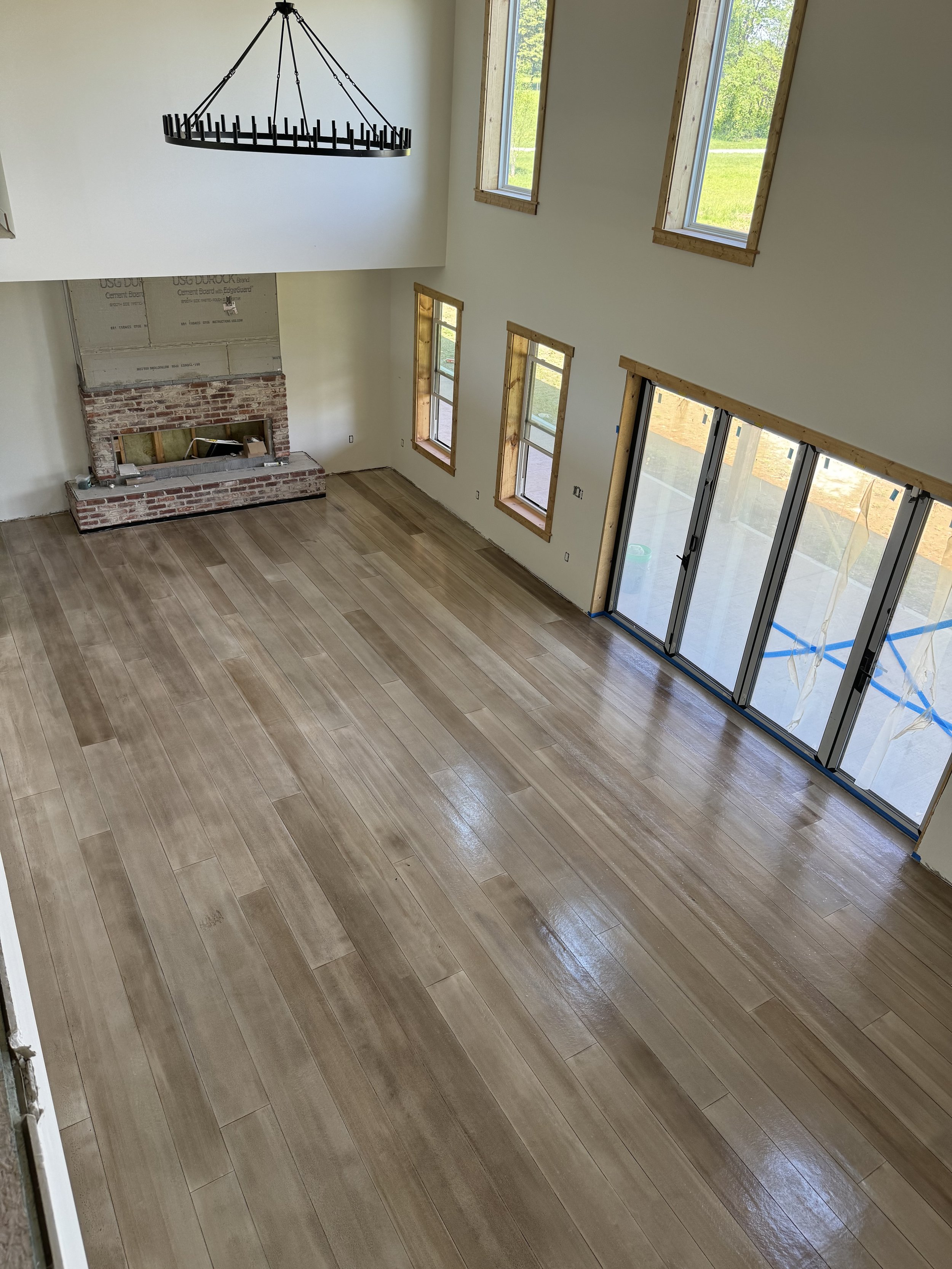 Interior of a house under construction with a spacious living room, hardwood flooring, a brick fireplace, and multiple windows and sliding glass doors awaiting finishing touches.