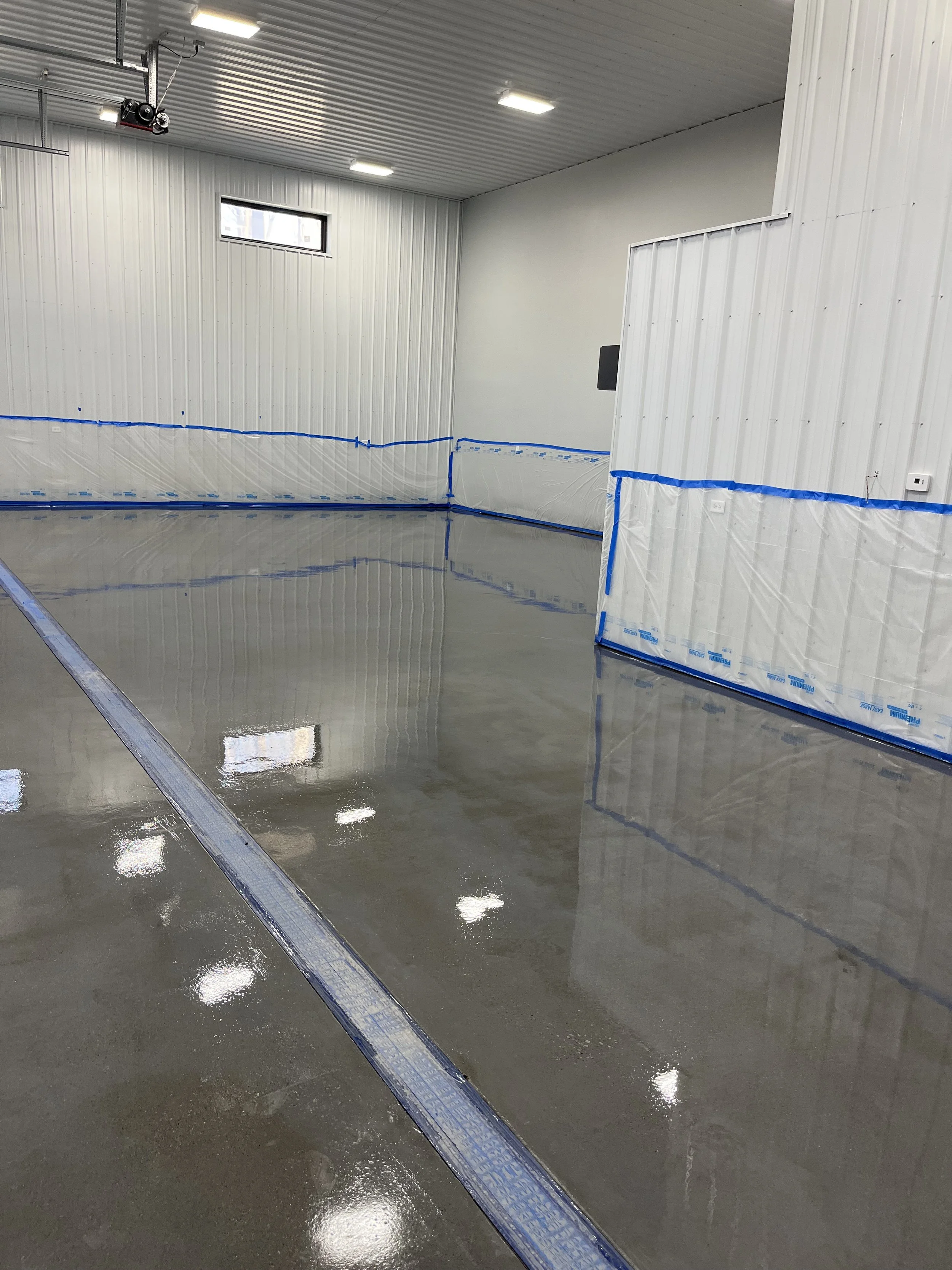 An indoor room with white corrugated metal walls and ceiling, small rectangular window, bright ceiling lights, and a shiny concrete floor with a drain channel, likely a preparation for sealing or coating.