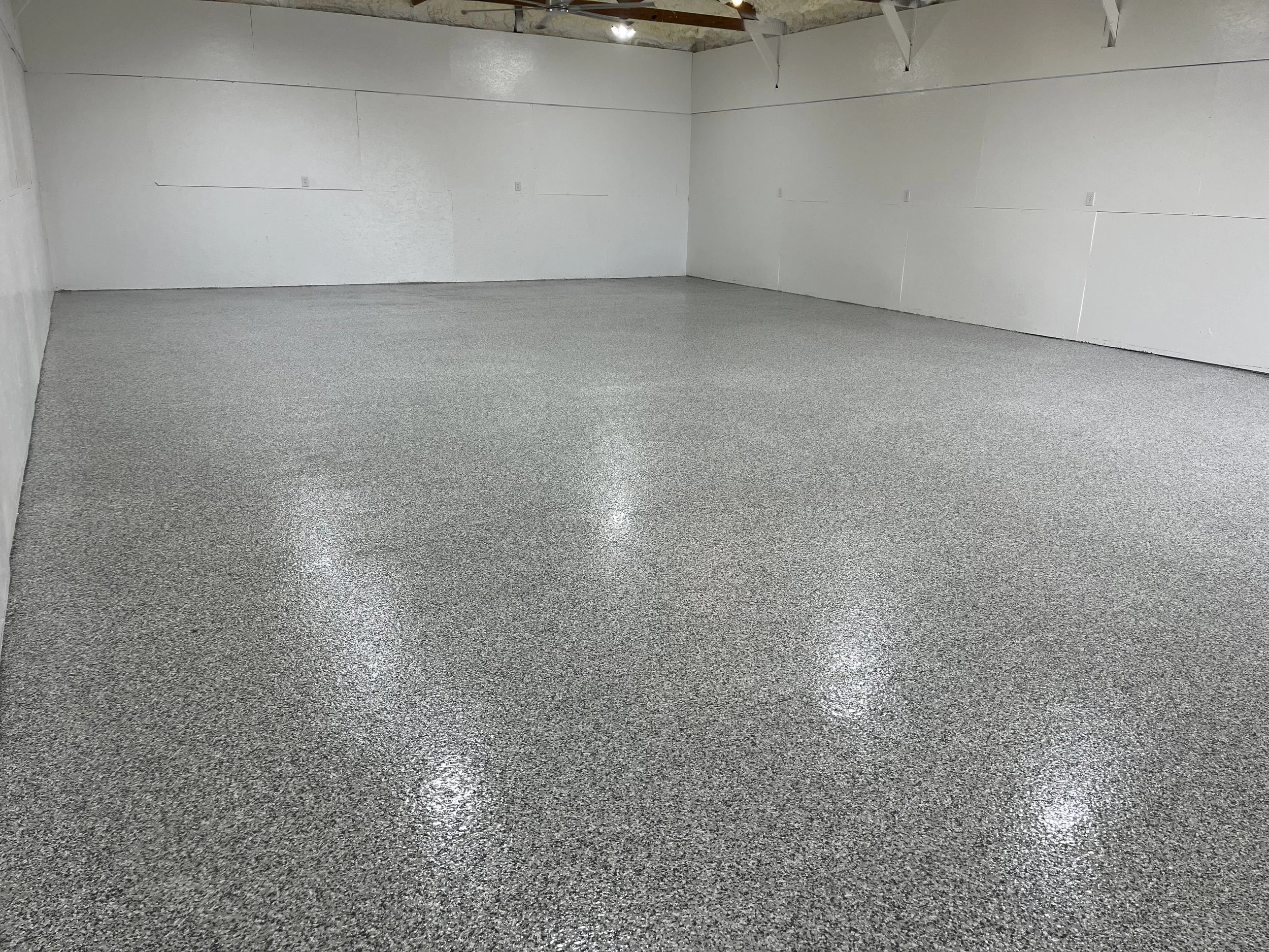 Empty garage with gray speckled epoxy floor, white walls, and ceiling with exposed wooden beams.