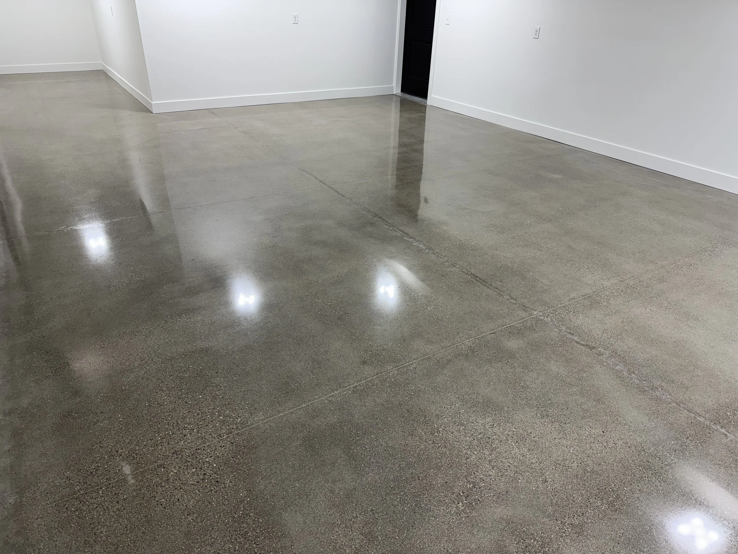 Smooth, polished concrete floor in a modern interior with white walls and baseboards, reflecting light from overhead fixtures.