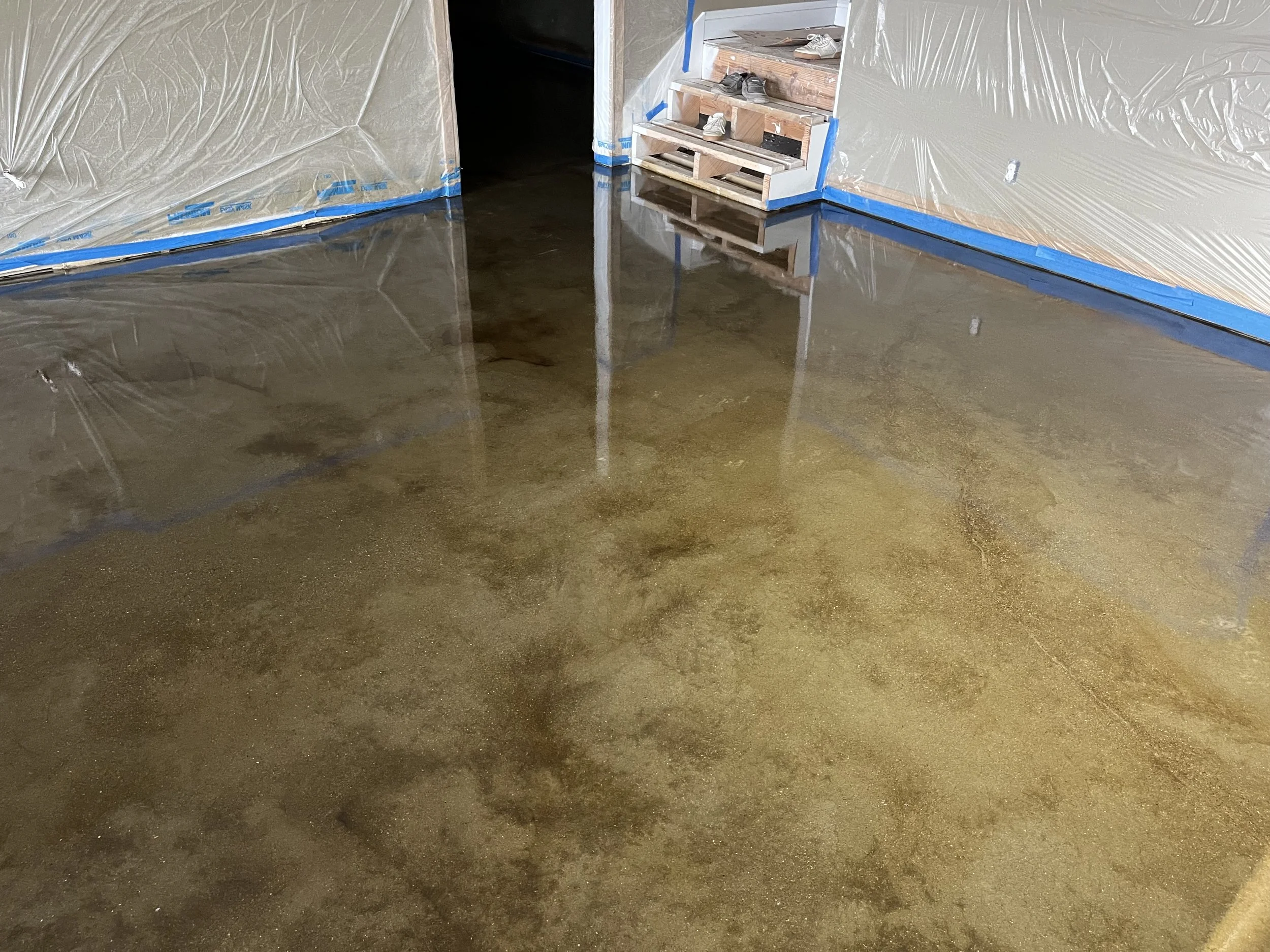 A room with a freshly poured concrete floor, plastic sheeting covering the walls, and a small shoe rack with sneakers at the far end.