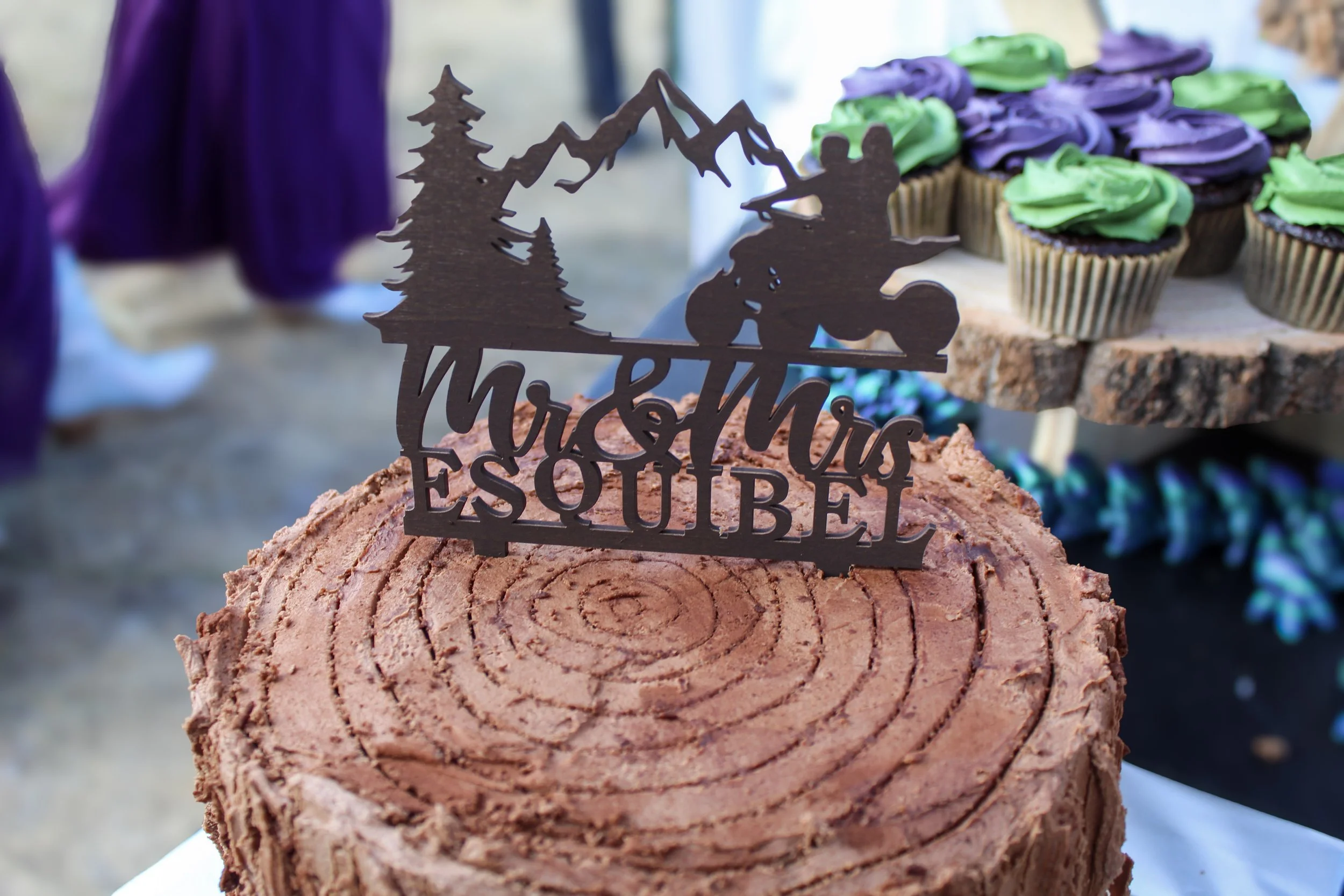 Chocolate cake with a wood grain pattern and a 'Mush Muskeg Esquibel' themed cake topper, with some cupcakes decorated with purple and green frosting in the background.