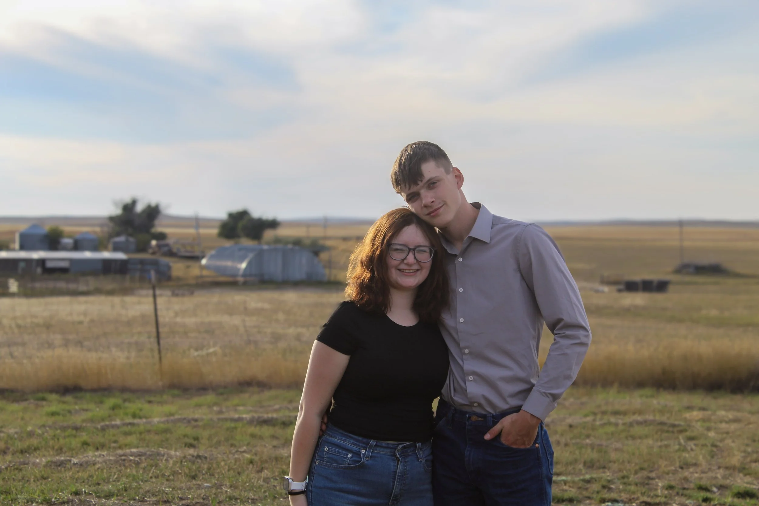 A young man and woman embrace outdoors in a field, smiling at the camera with farm structures in the background and a partly cloudy sky.