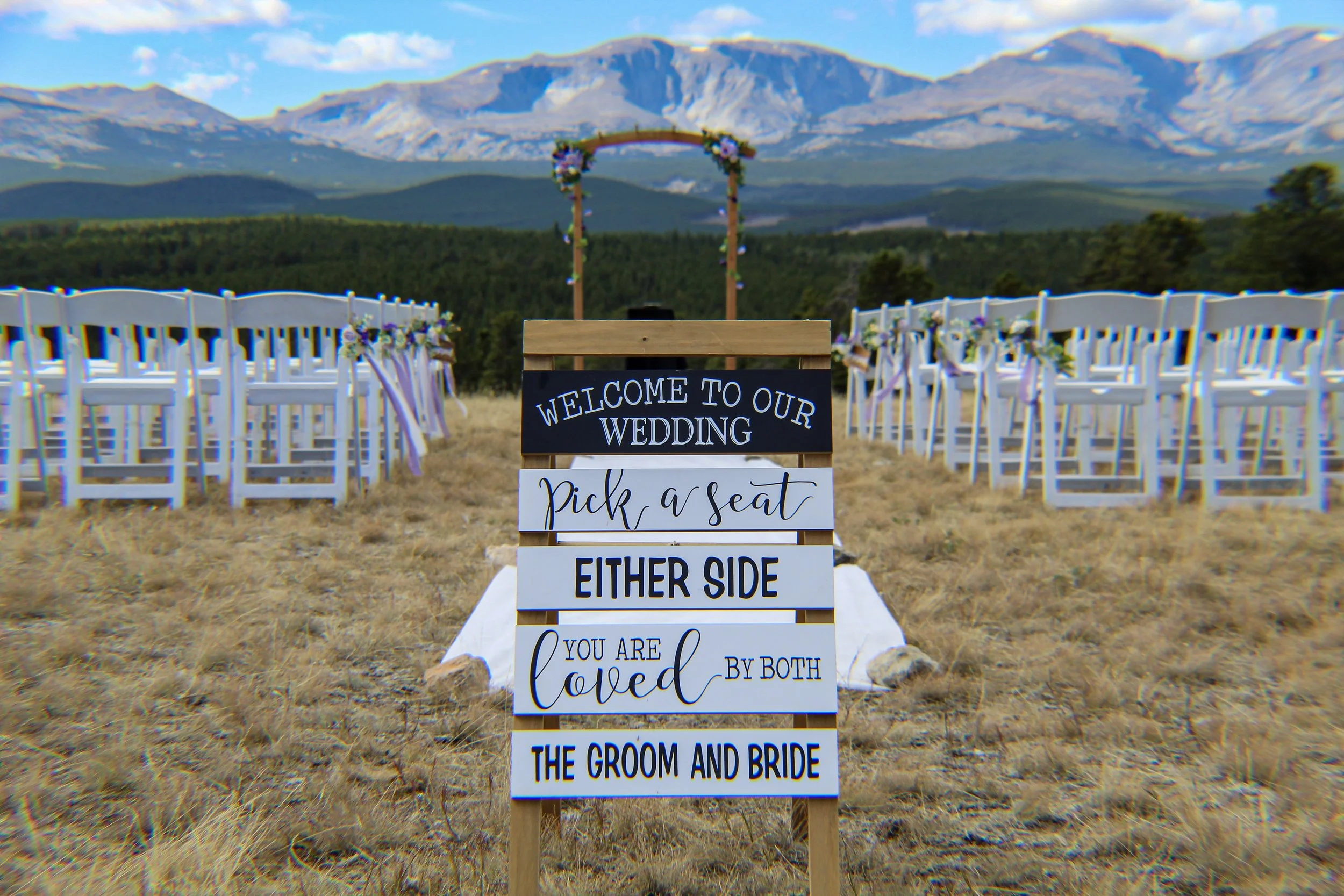 Wedding venue setup outdoors with white chairs decorated with flowers, an arch in the background, mountainous landscape, and a sign that says "Welcome to our wedding, pick a seat, either side, you are loved by both the groom and bride."