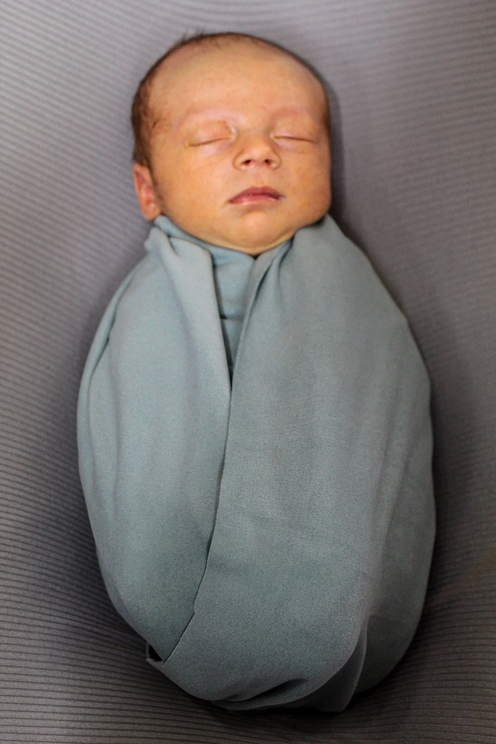 Swaddled newborn baby sleeping with eyes closed, wrapped in a light gray blanket on a dark gray surface.